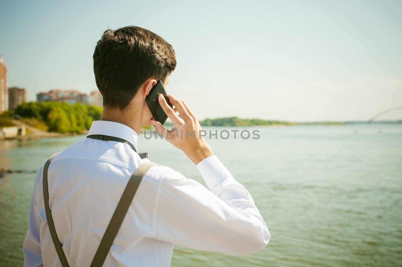 young skinny man, elegantly stylishly dressed in white shirt, gray trousers with suspenders and bow tie. Portrait of young guy talking on phone on background of river on warm sunny summer day