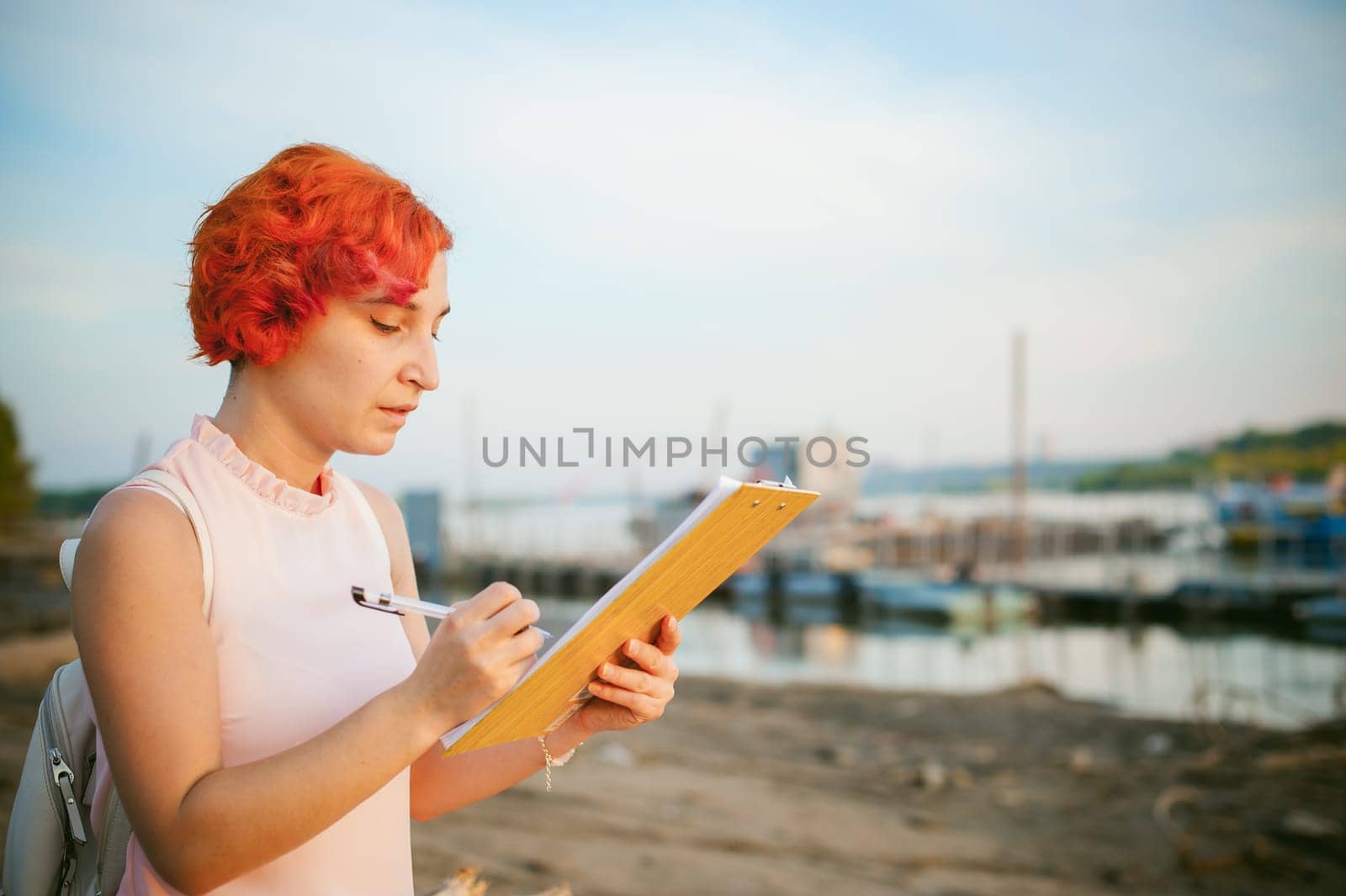 woman with dyed red hair in a pale pink dress with white backpack, signed important documents sitting outdoors near the river at sunset, against a boat mooring