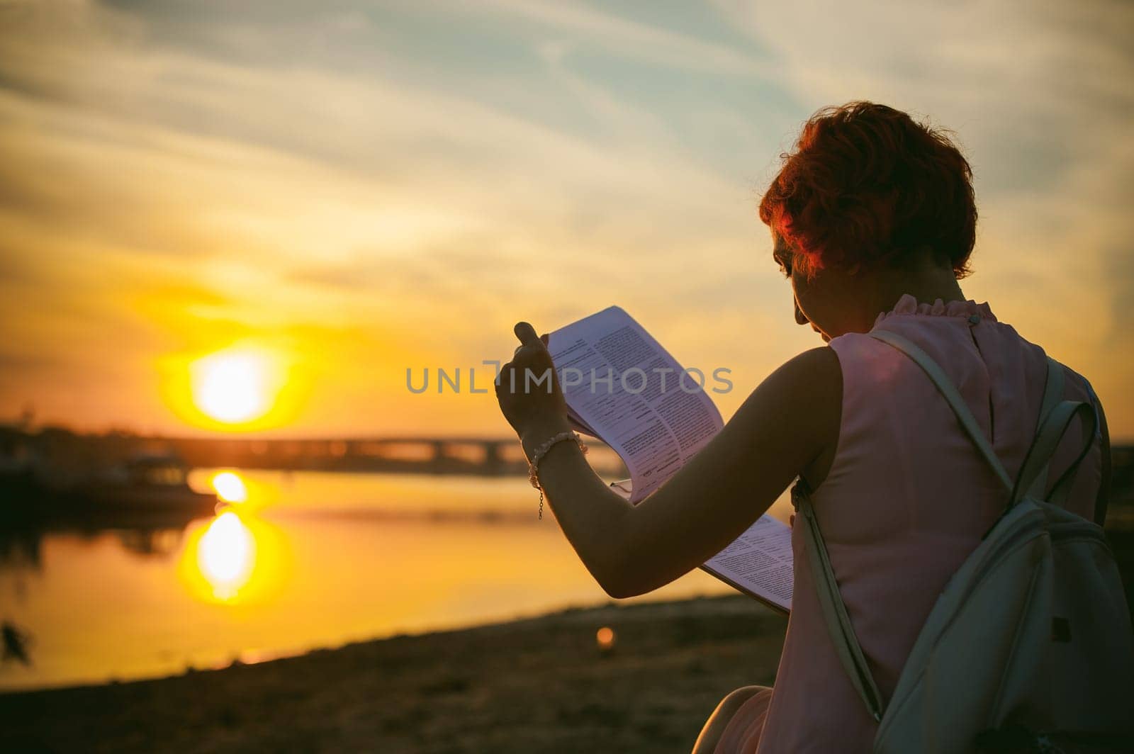woman with dyed red hair in a pale pink dress with white backpack, signed important documents sitting outdoors near the river at sunset, against a boat mooring