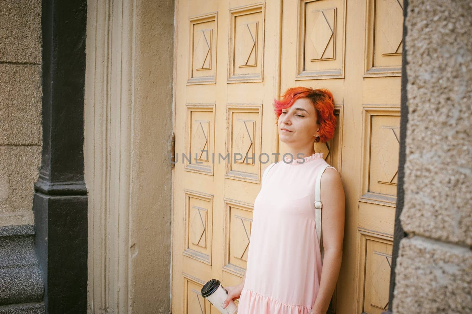 drinks coffee from a cup. woman with dyed red hair in a pale pink dress with a backpack white color drinking coffee, holding a paper cup of Take away, on a city street a warm summer day, behind, a huge exclusive doors