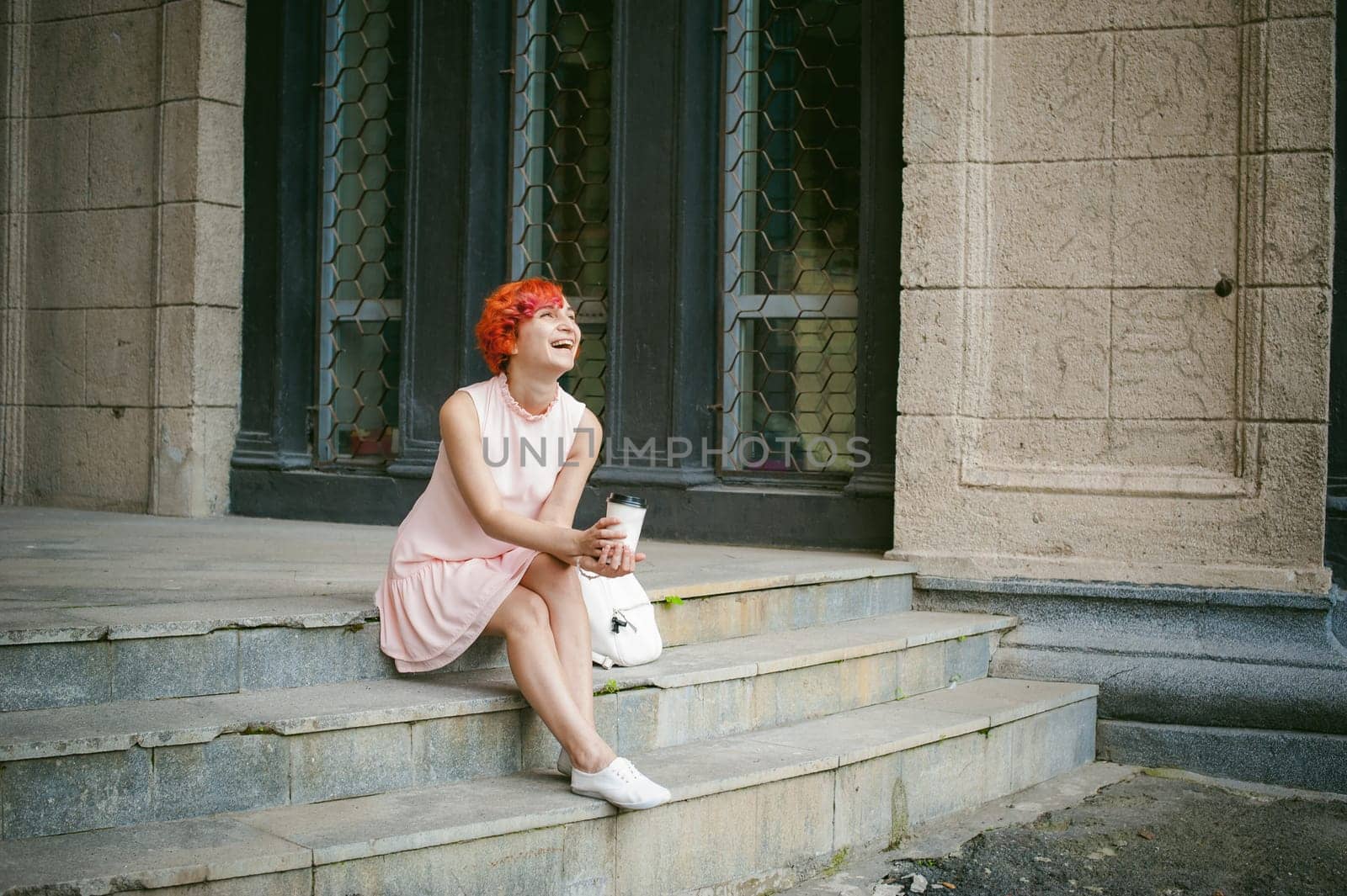 drinks coffee from a cup. woman with dyed red hair in a pale pink dress with a backpack white color drinking coffee, holding a paper cup of Take away, on a city street a warm summer day