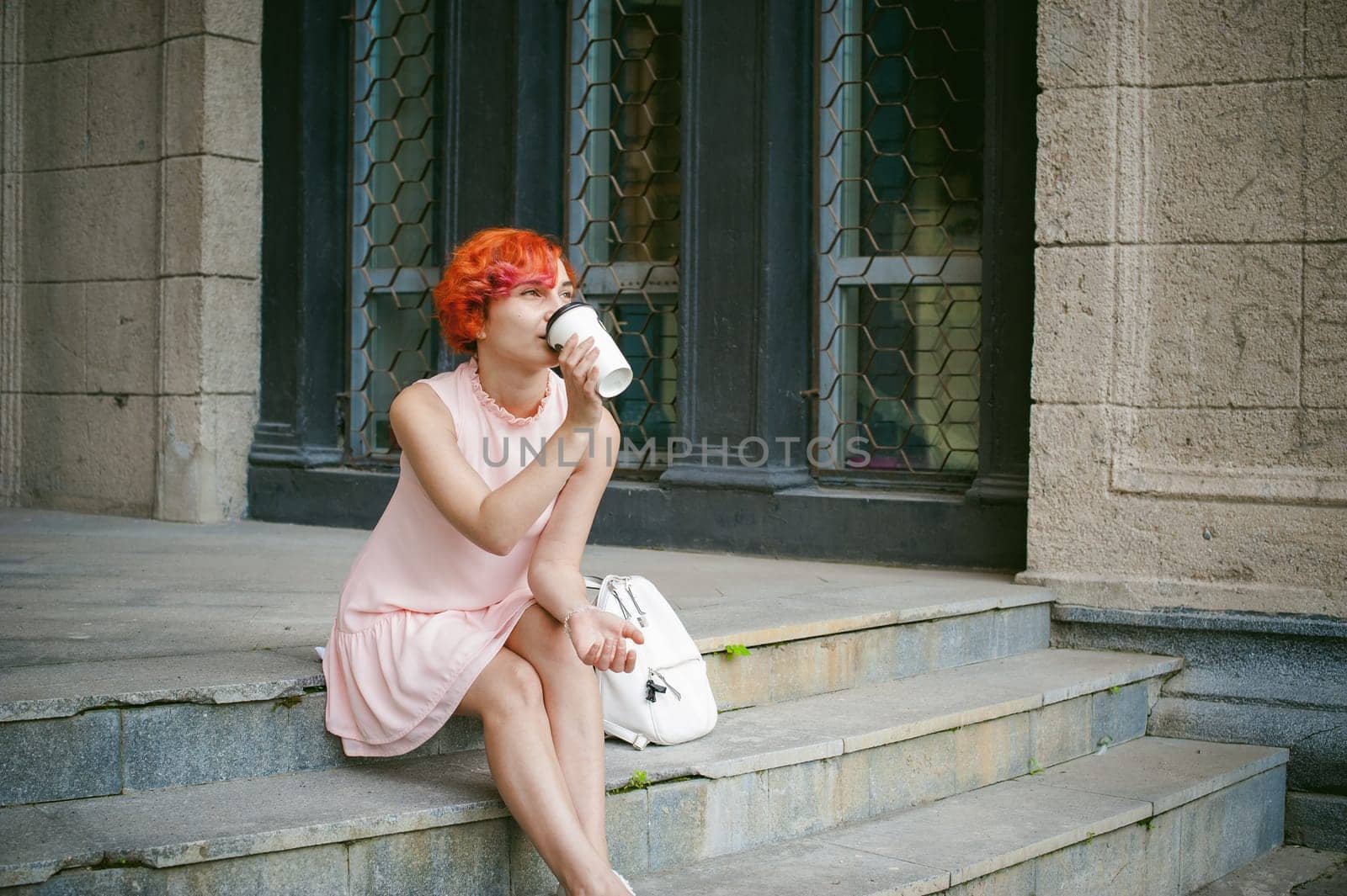 drinks coffee from a cup. woman with dyed red hair in a pale pink dress drinking coffee, holding a paper cup of coffee to go, sitting on the steps outside a summer day.