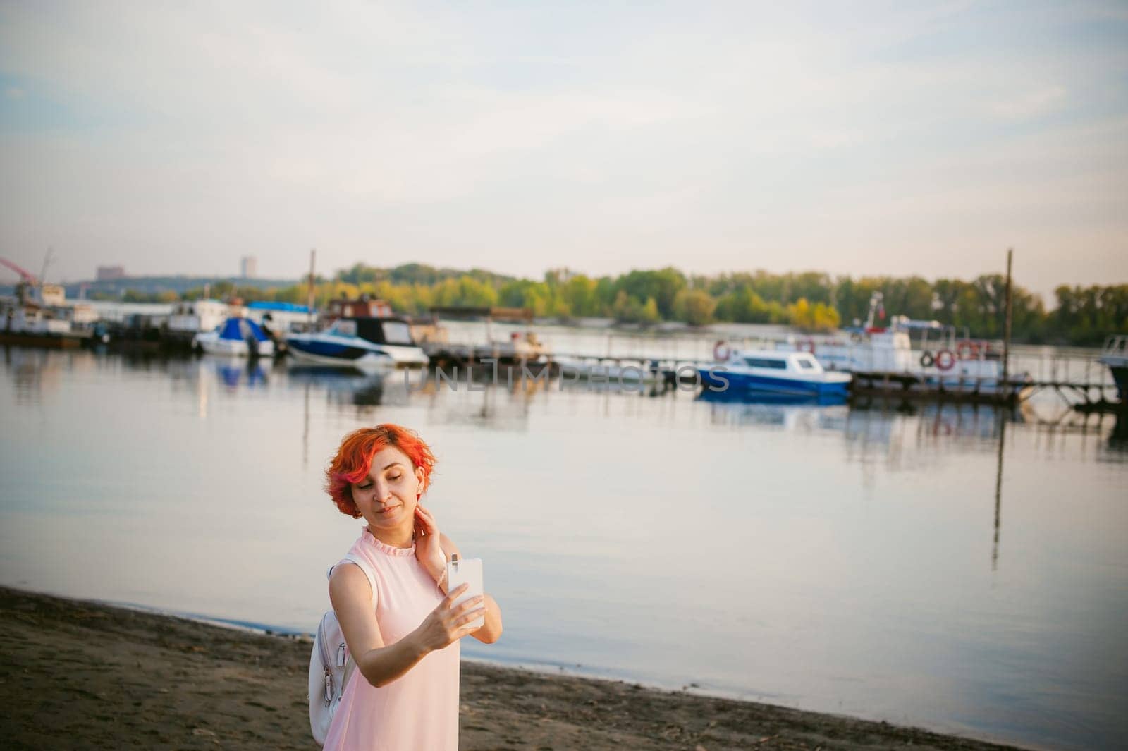 girl in pale pink dress with red hair and backpack walking along river bank, pictures of themselves on their mobile camera phone, against backdrop of boats moored on a warm summer day