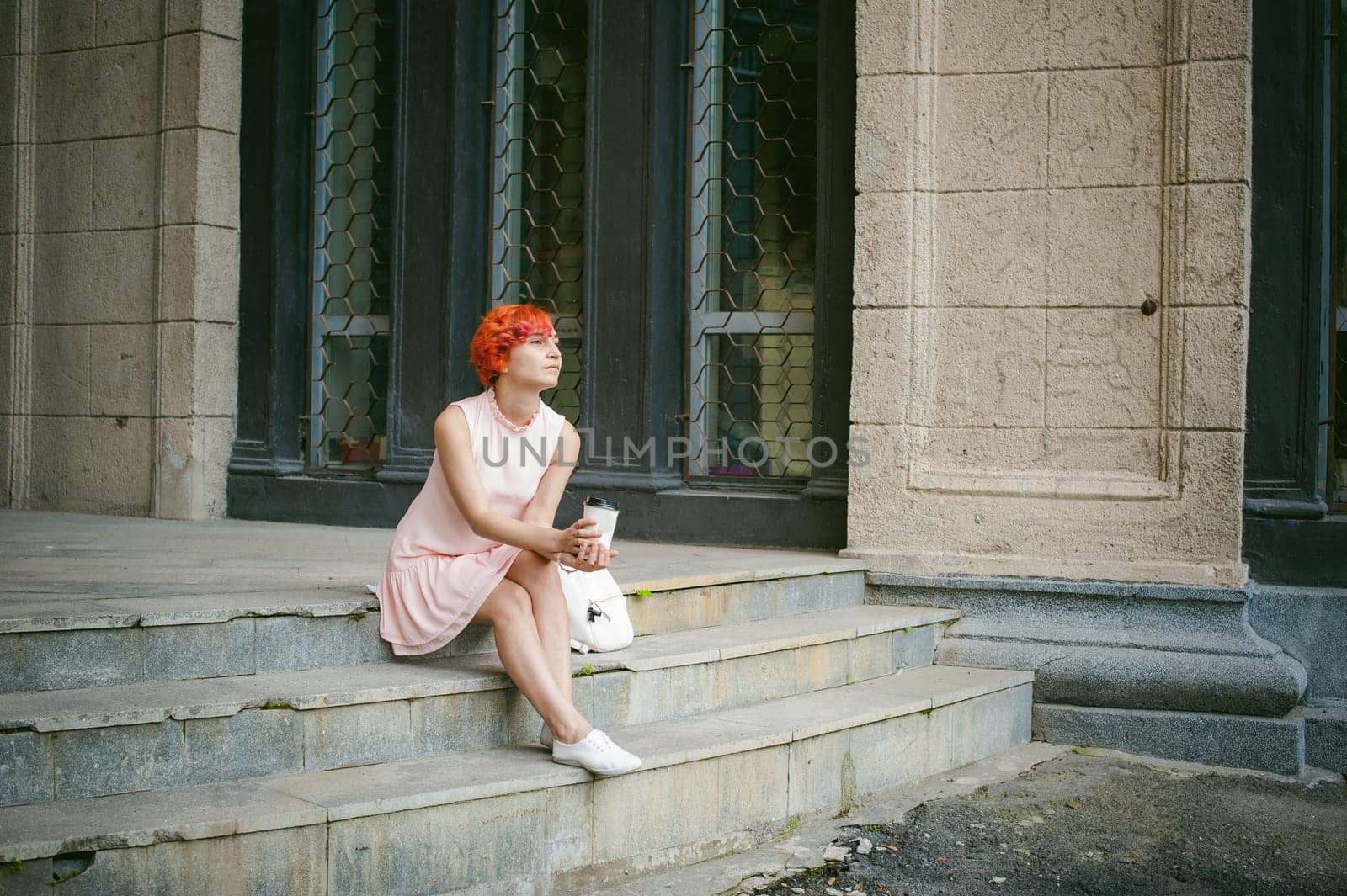 drinks coffee from a cup. woman with dyed red hair in a pale pink dress with a backpack white color drinking coffee, holding a paper cup of Take away, on a city street a warm summer day