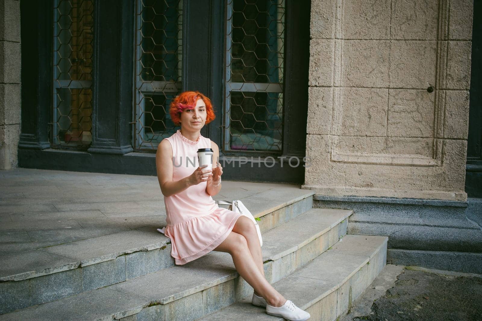 drinks coffee from a cup. woman with dyed red hair in a pale pink dress drinking coffee, holding a paper cup of coffee to go, sitting on the steps outside a summer day.