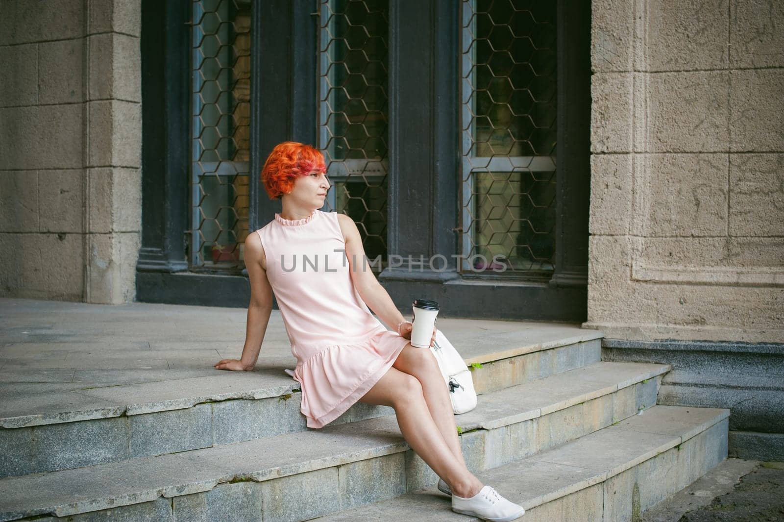 drinks coffee from a cup. woman with dyed red hair in a pale pink dress drinking coffee, holding a paper cup of coffee to go, sitting on the steps outside a summer day.