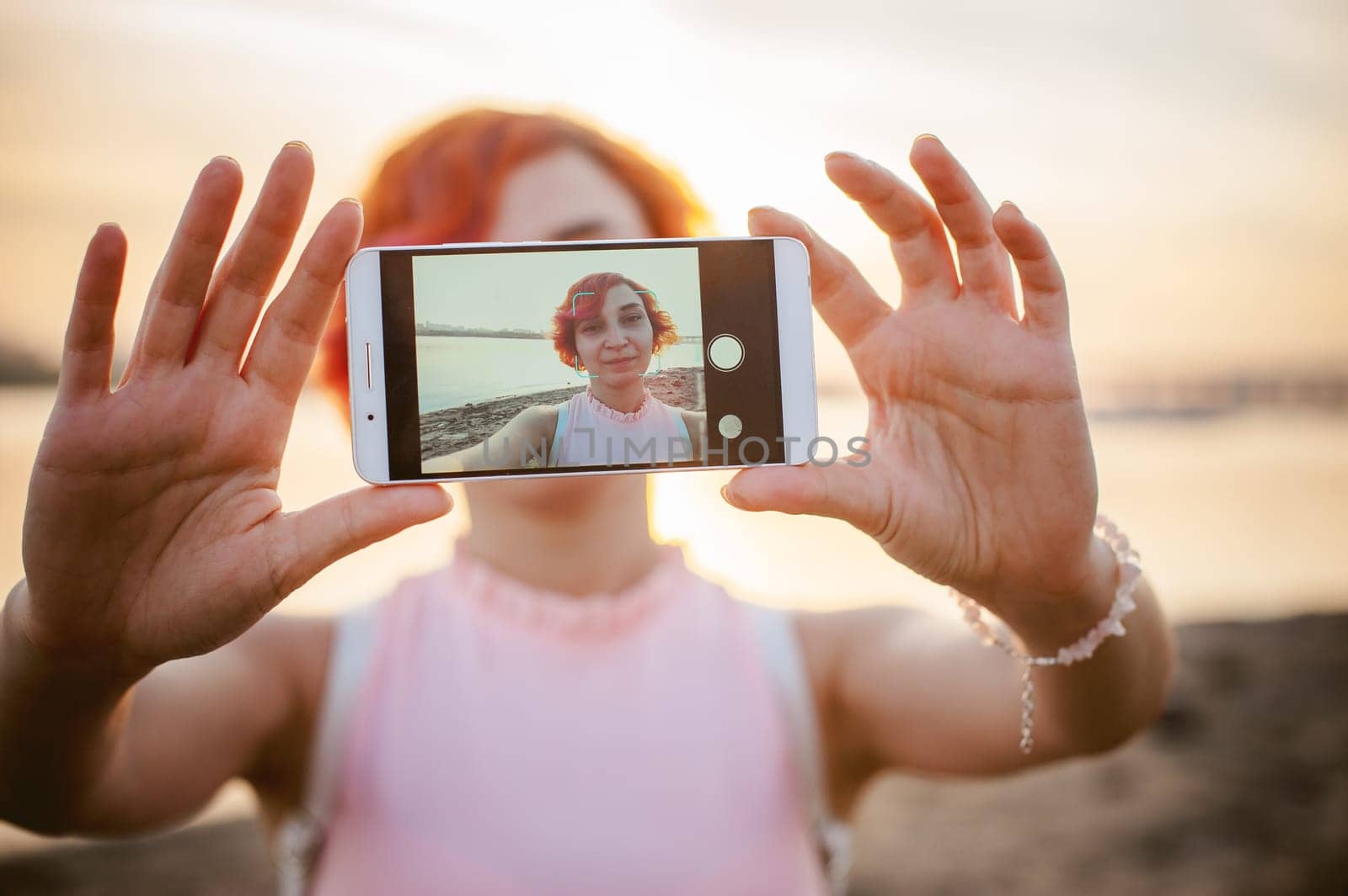 girl in pale pink dress with red hair and backpack walking along river bank, pictures of themselves on their mobile camera phone, against backdrop of boats moored on a warm summer day