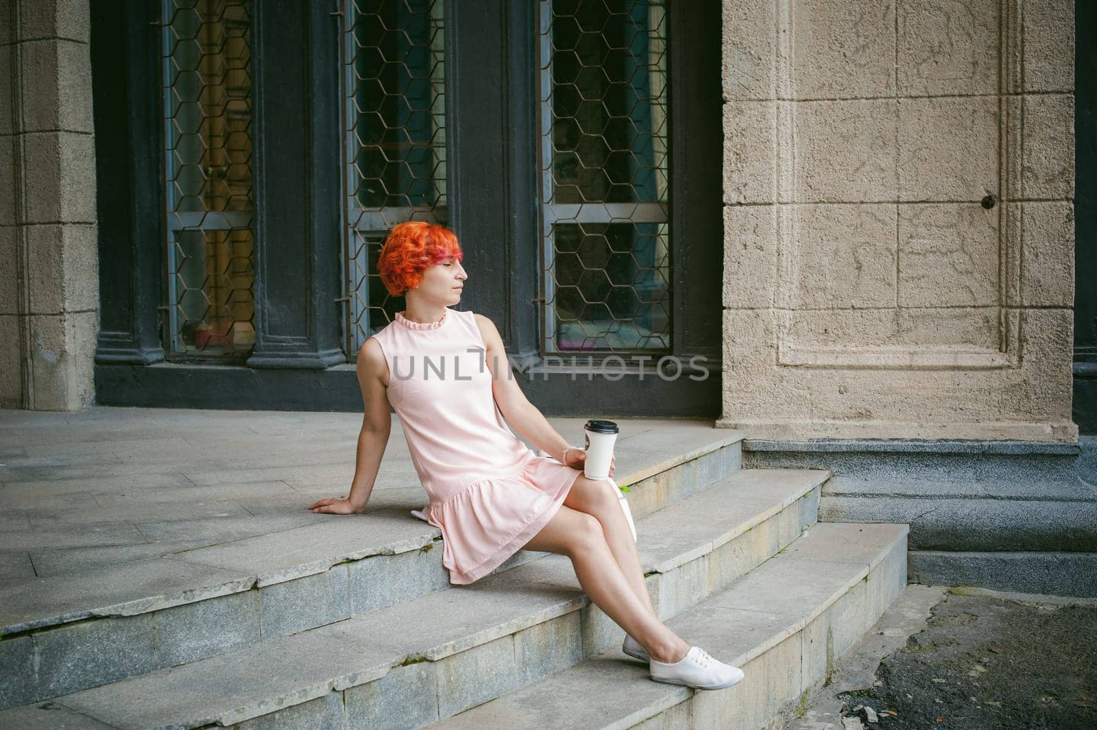 drinks coffee from a cup. woman with dyed red hair in a pale pink dress drinking coffee, holding a paper cup of coffee to go, sitting on the steps outside a summer day.