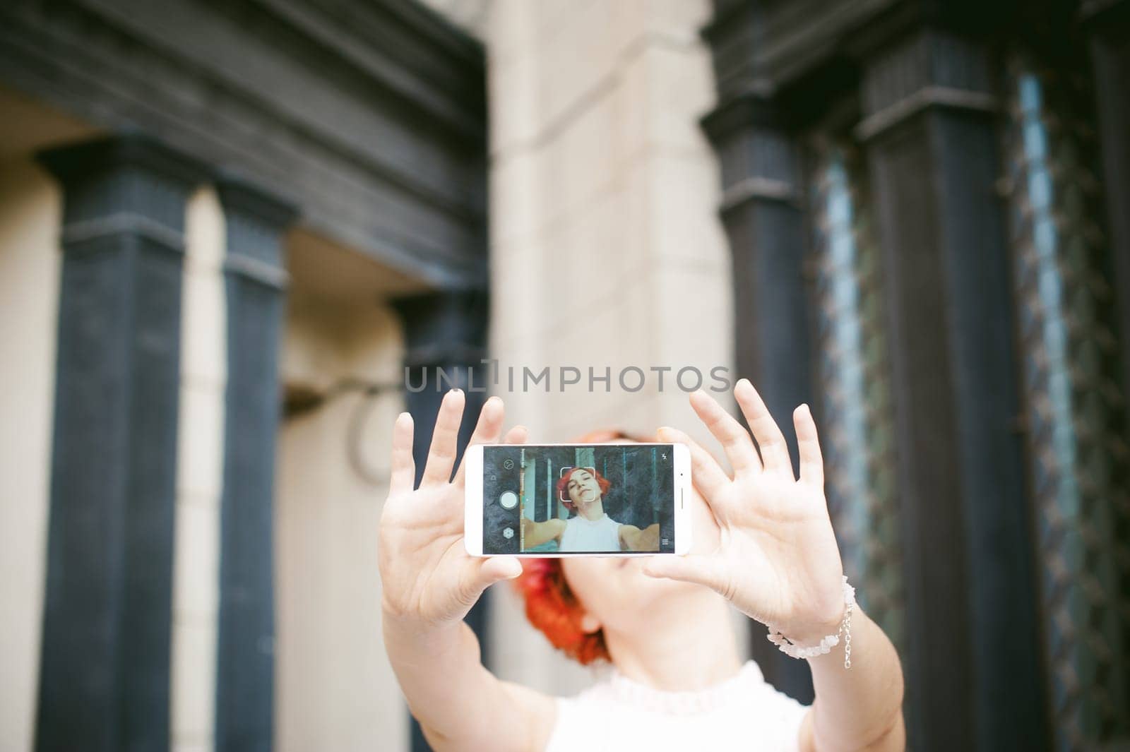 portrait of a girl doing selfie. girl in light pink dress with red dyed hair, pictures of themselves on their mobile camera phone, sitting on the stairs summer day