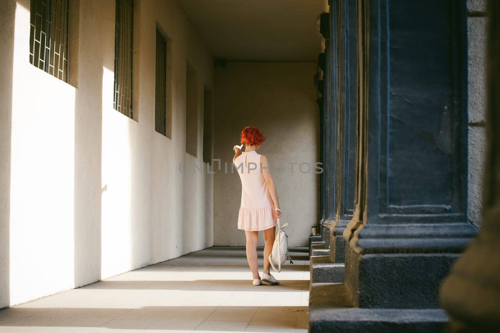 girl in a pale pink dress with dyed red hair, a white backpack in hand, walking between light and shadow on the columns sunny summer day