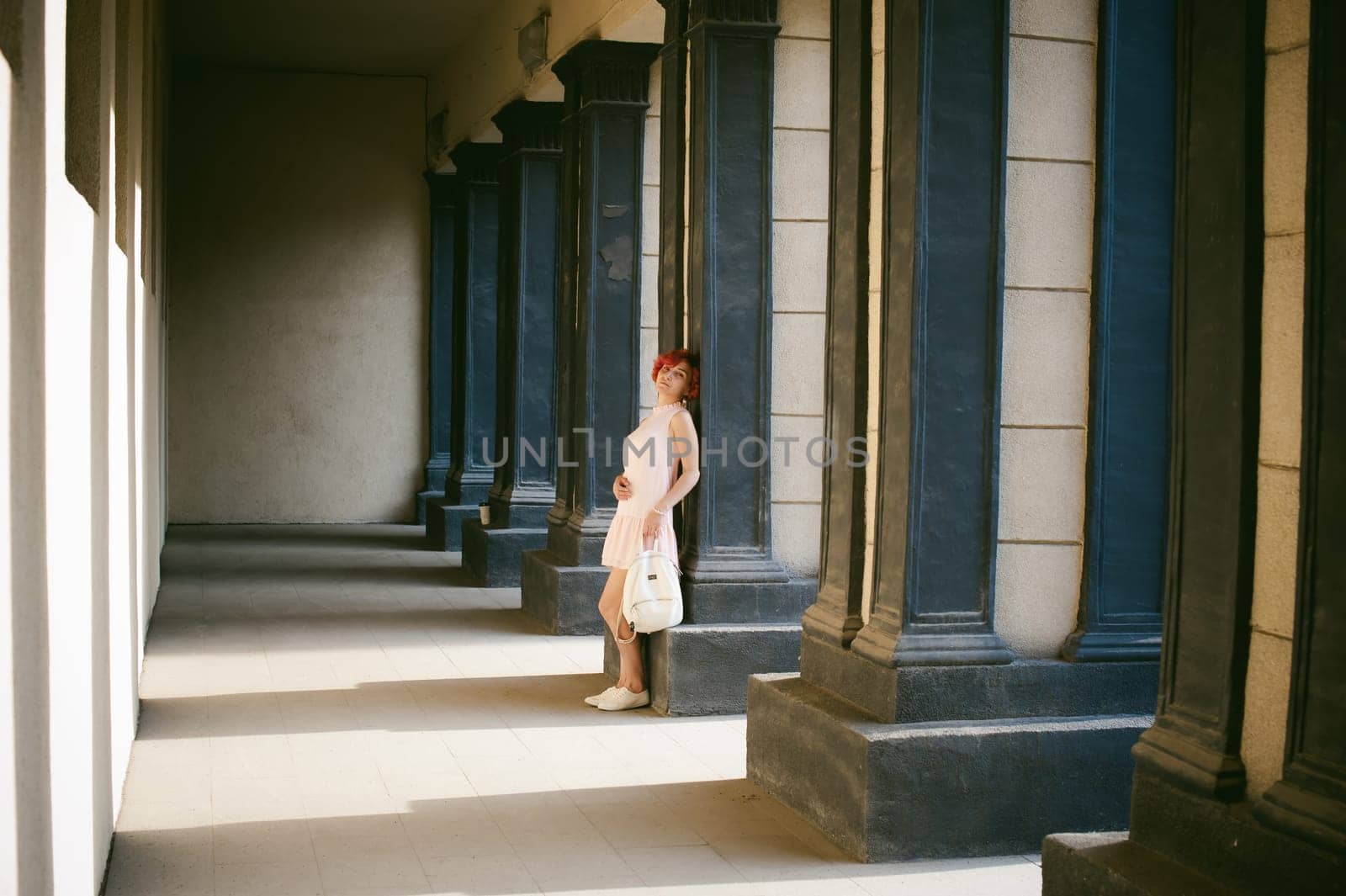 girl in a pale pink dress with dyed red hair, a white backpack in hand, walking between light and shadow on the columns sunny summer day