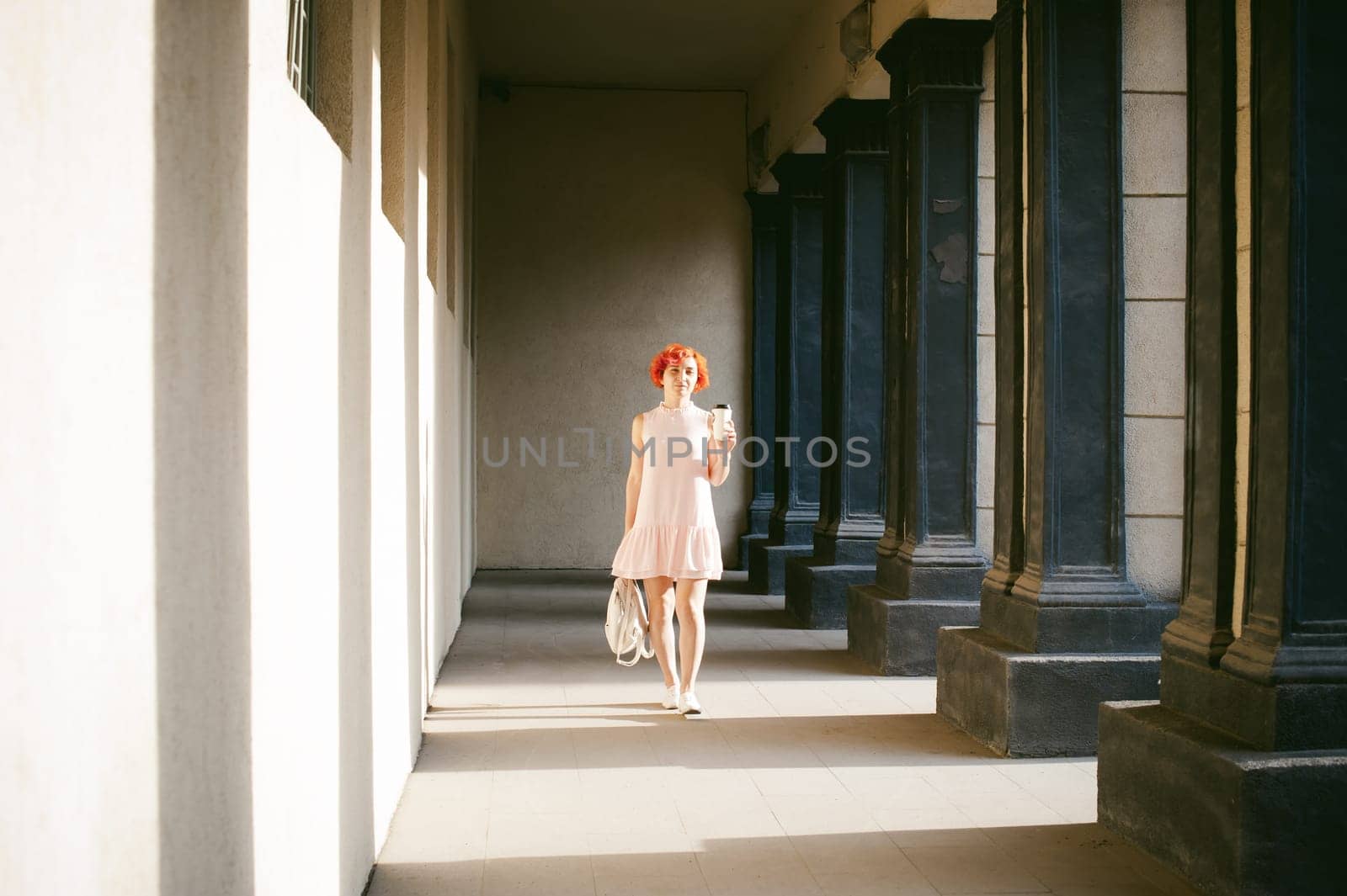 girl in a pale pink dress with dyed red hair, a white backpack in hand, drinks coffee from a cup, walking between light and shadow on the columns sunny summer day