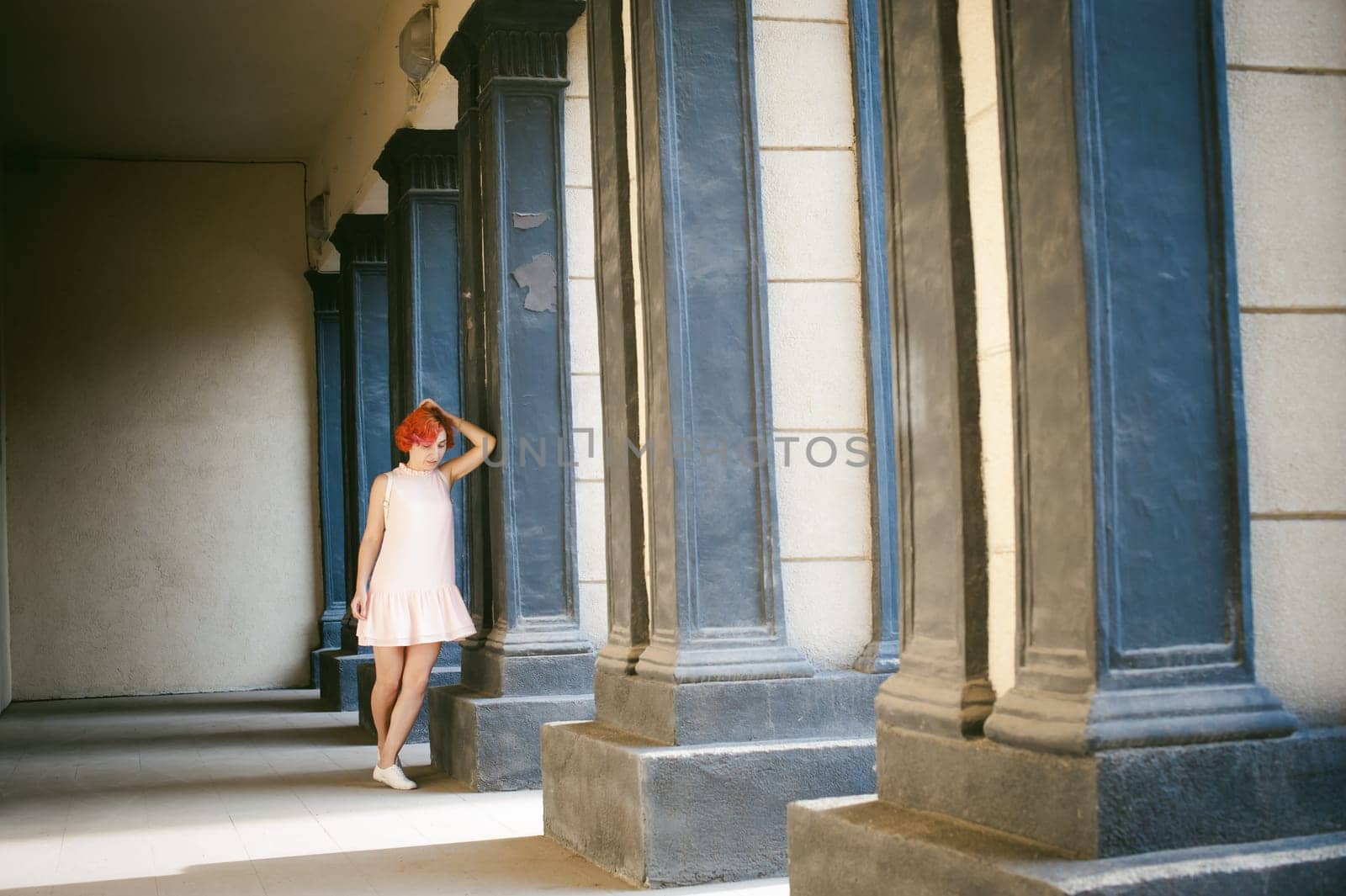 girl in a pale pink dress with dyed red hair, a white backpack in hand, walking between light and shadow on the columns sunny summer day