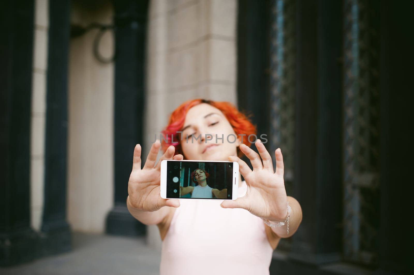 portrait of a girl doing selfie. girl in light pink dress with red dyed hair, pictures of themselves on their mobile camera phone, sitting on the stairs summer day