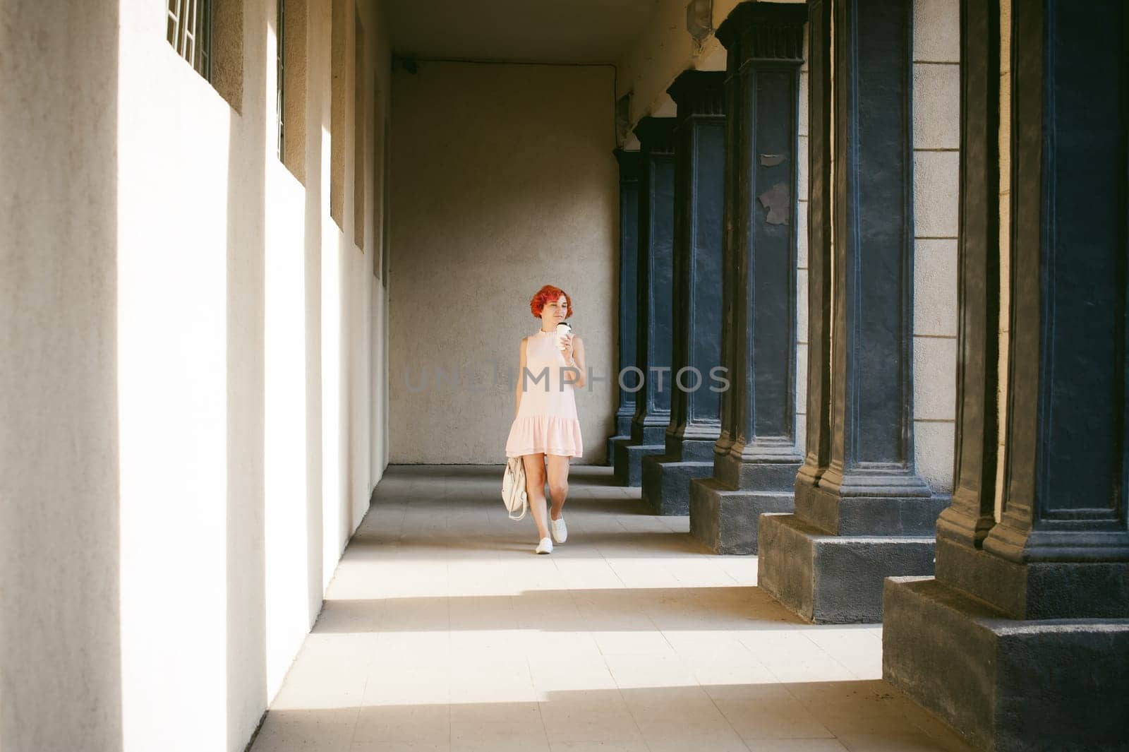 girl in a pale pink dress with dyed red hair, a white backpack in hand, drinks coffee from a cup, walking between light and shadow on the columns sunny summer day