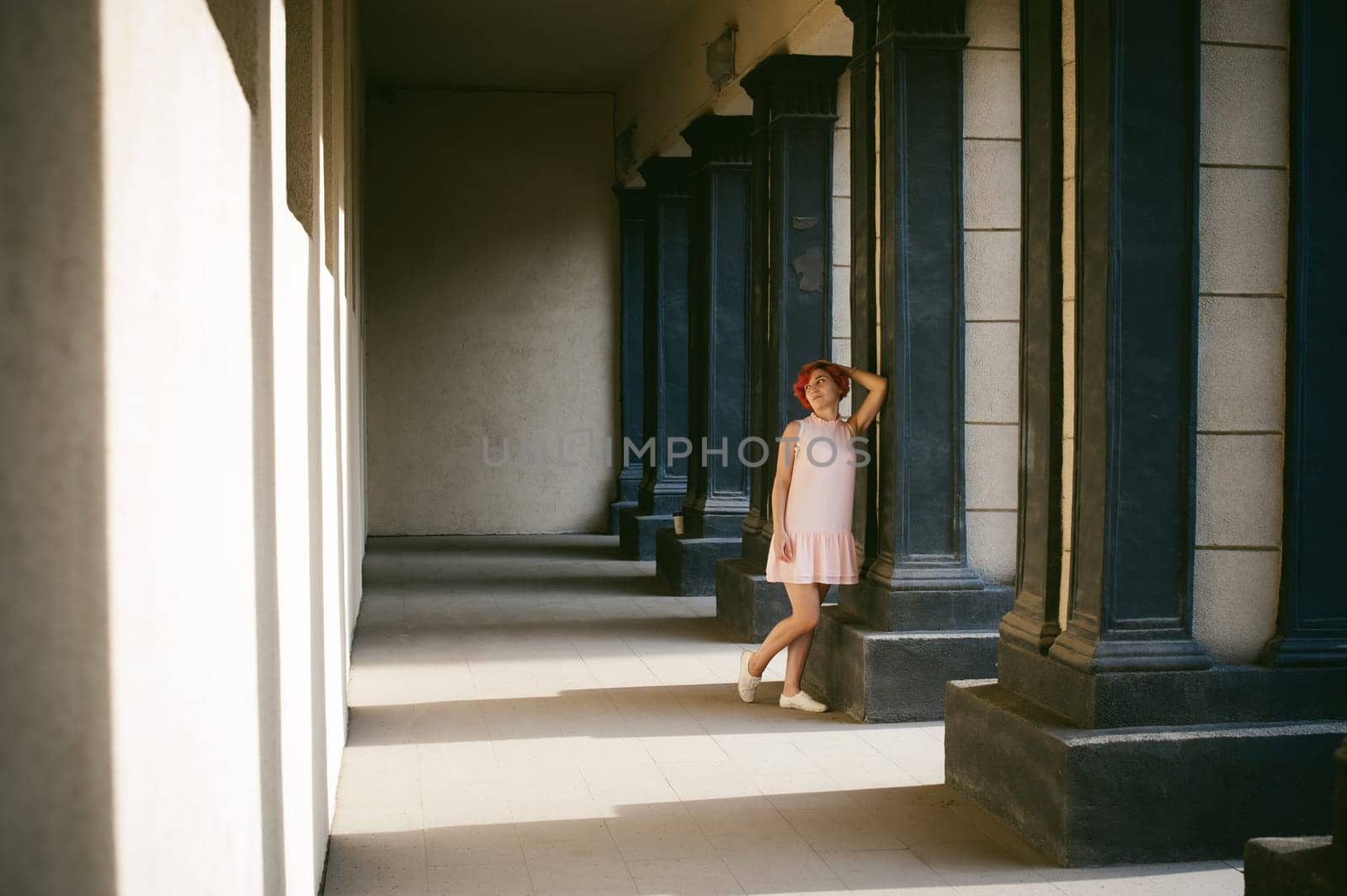 girl in a pale pink dress with dyed red hair, a white backpack in hand, walking between light and shadow on the columns sunny summer day