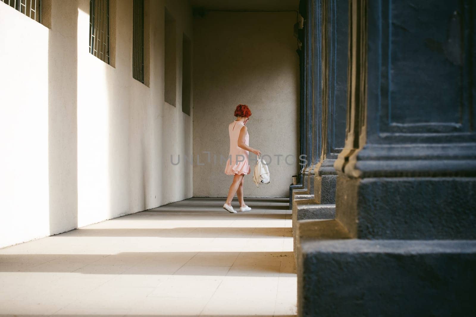 girl in a pale pink dress with dyed red hair, a white backpack in hand, walking between light and shadow on the columns sunny summer day