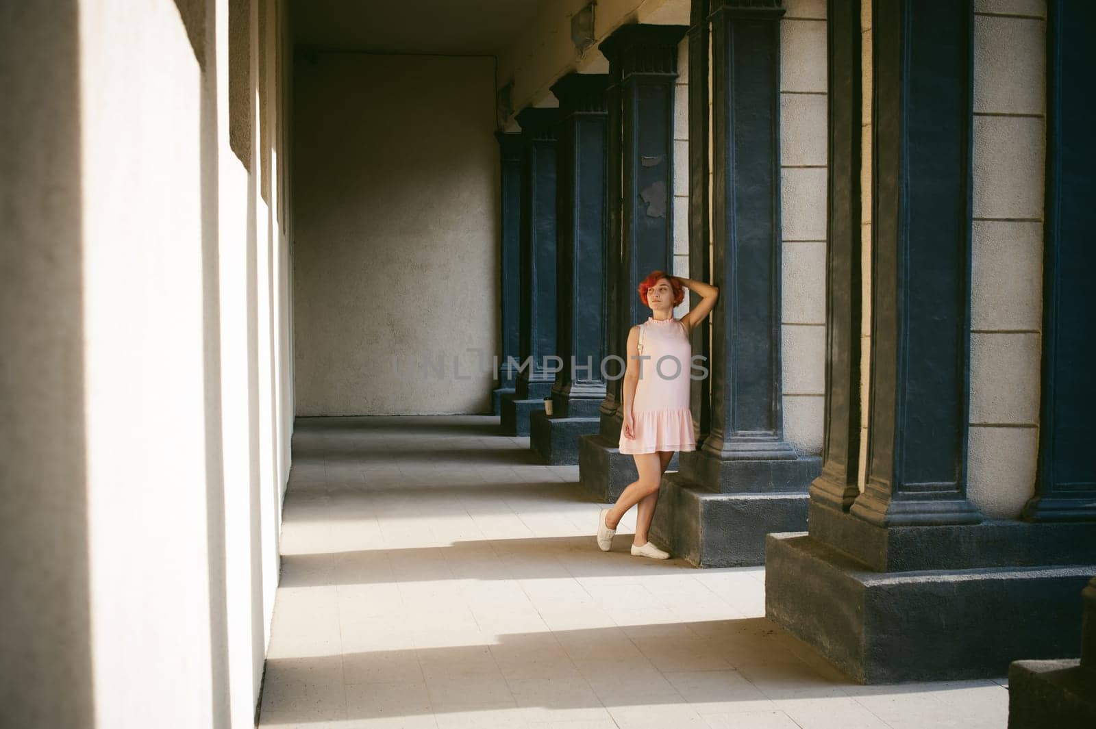 girl in a pale pink dress with dyed red hair, a white backpack in hand, walking between light and shadow on the columns sunny summer day