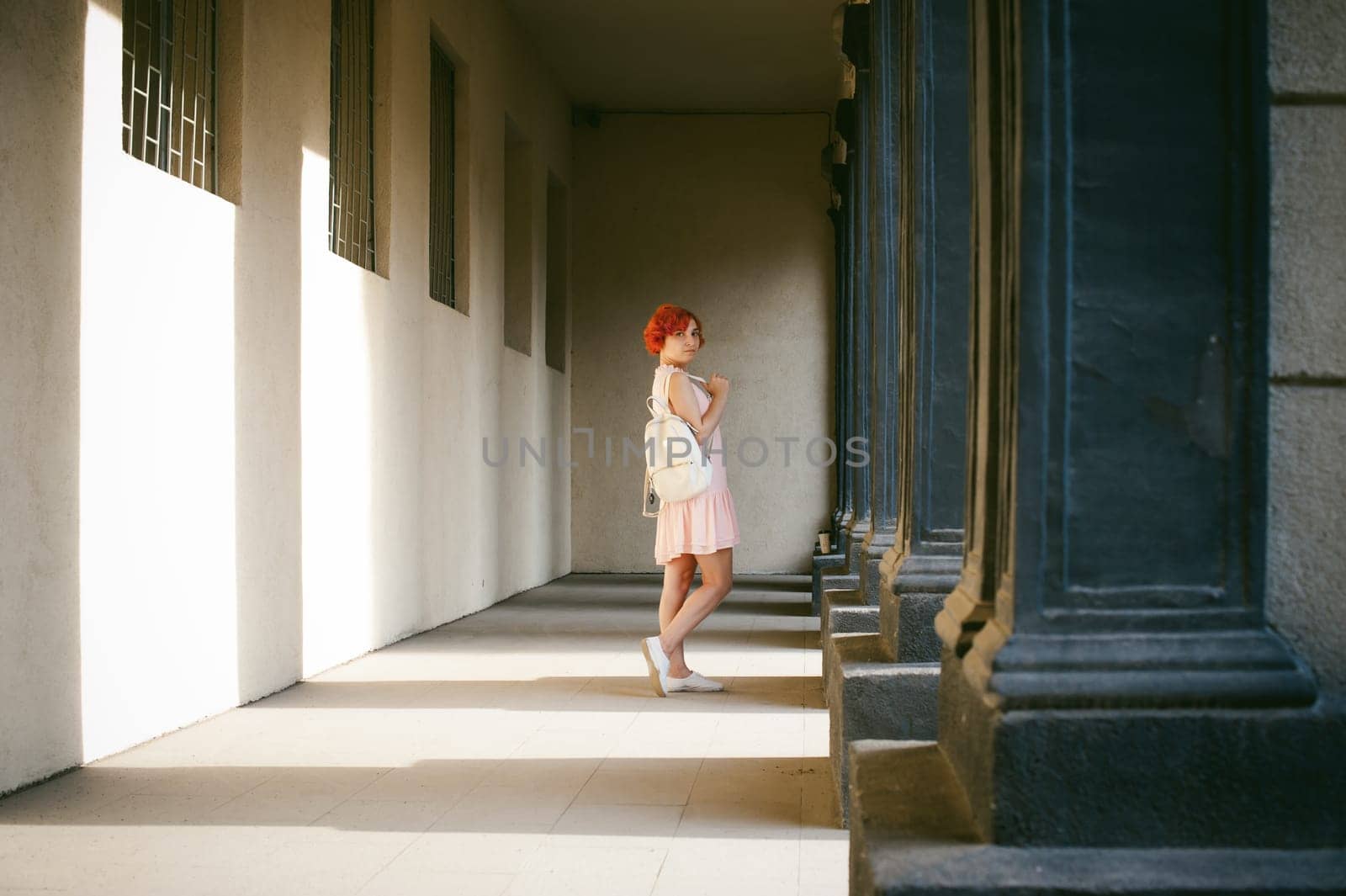 girl in a pale pink dress with dyed red hair, a white backpack in hand, walking between light and shadow on the columns sunny summer day