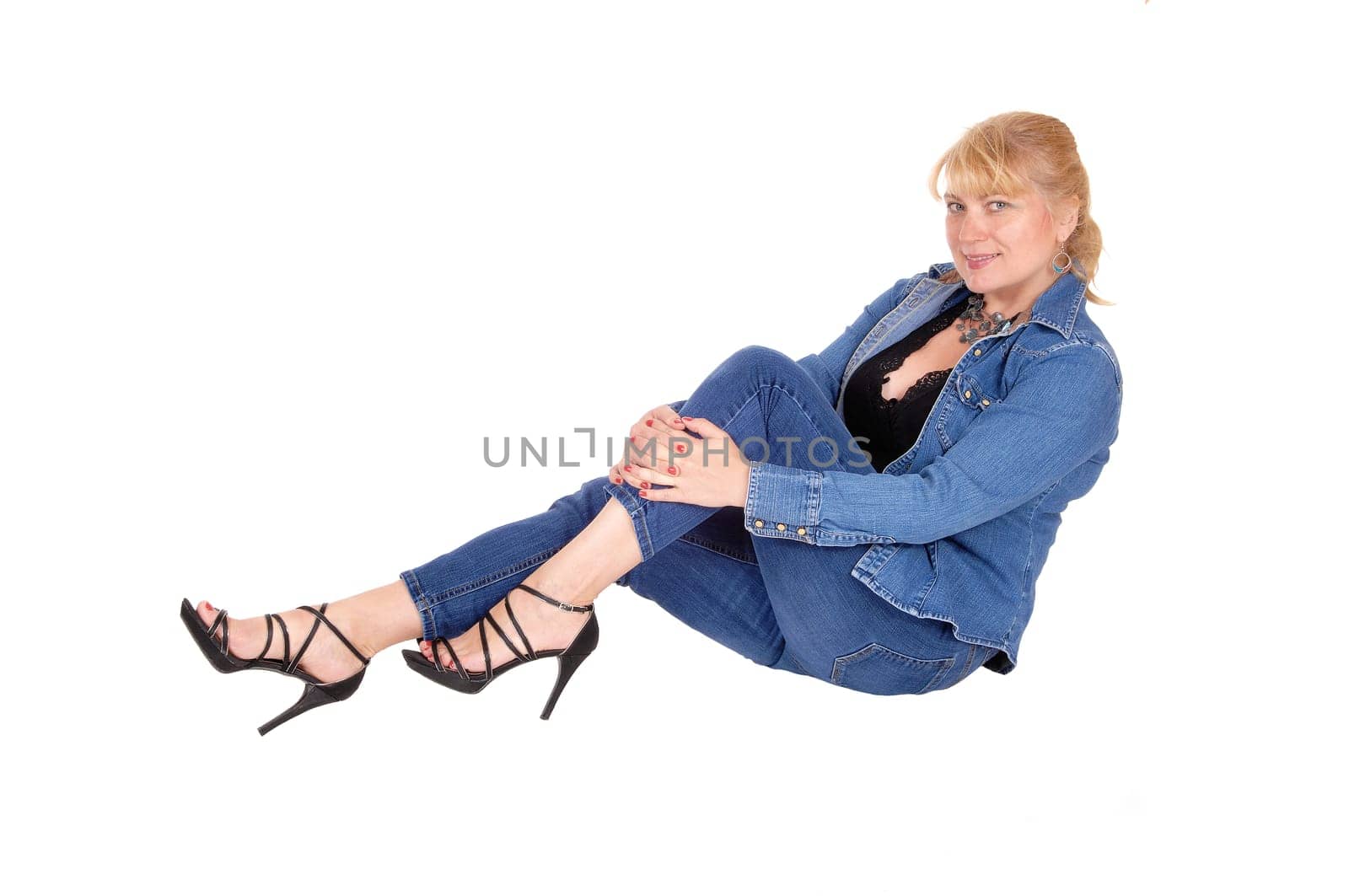 A lovely blond woman in a jeans outfit and high heels sitting on the.floor, smiling, isolated for white background..