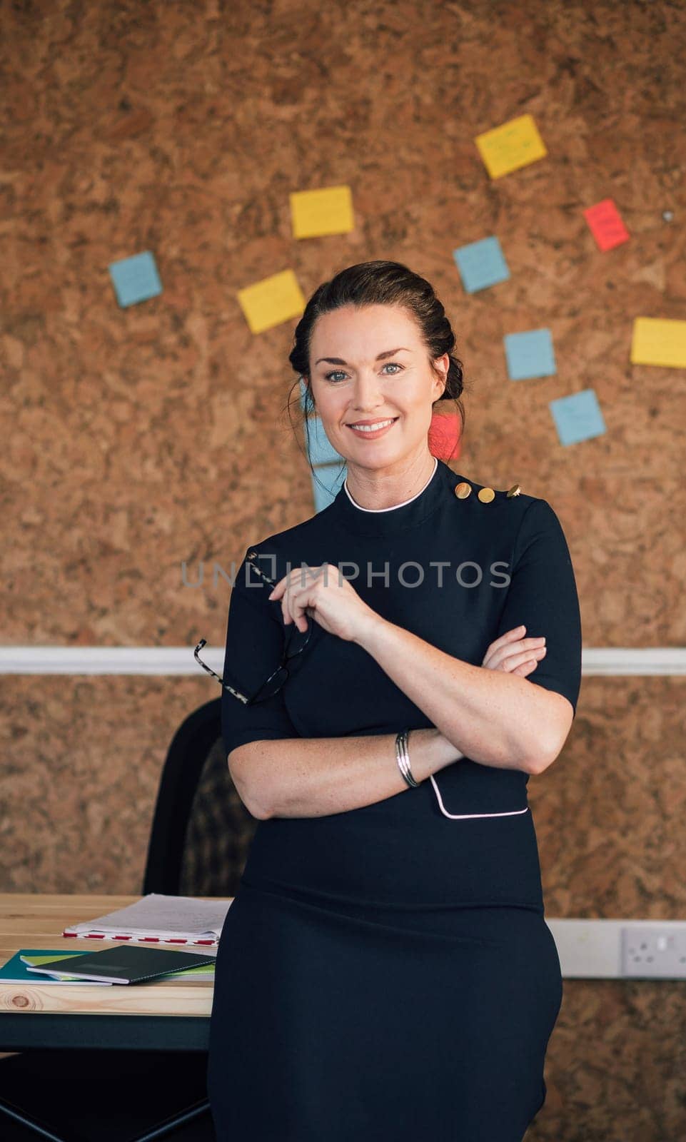 Portrait of a beautiful woman in her workplace. She is standing in front of a corkboard and is smartly dressed, smiling for the camera.