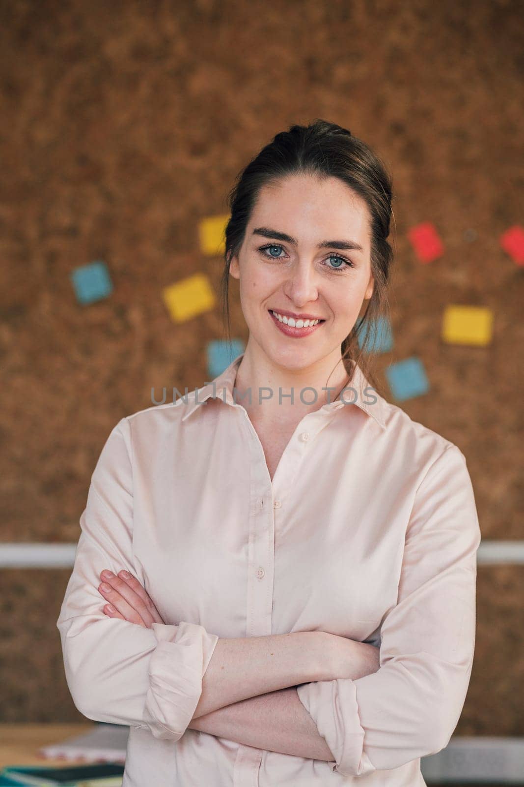 Portrait of a female office worker. She is standing in front of a cork board, smartly dressed with her arms folded and is smiling for the camera.