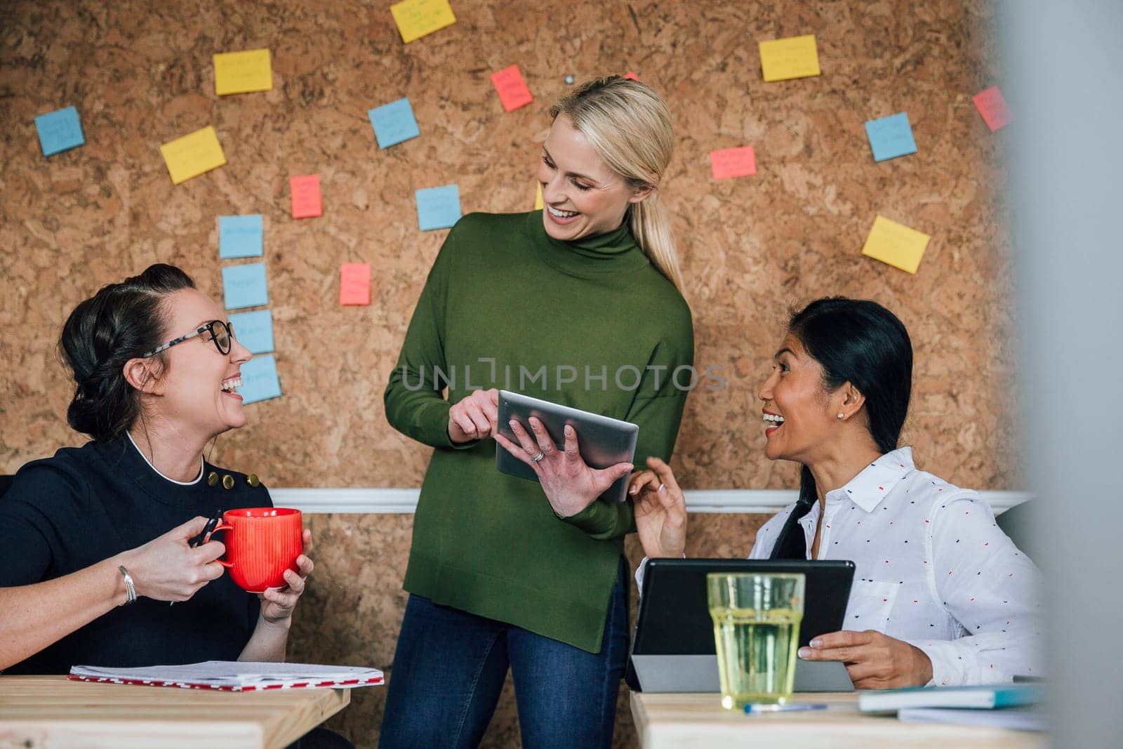 Women are working together in an office. They are talking and laughing as they use digital tablets.
