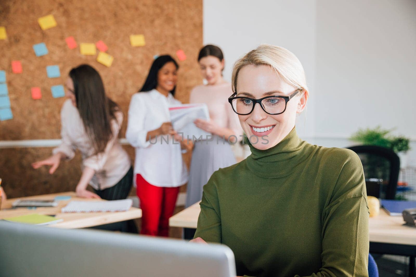 Beautiful woman is smiling while working at her desk in her workplace. There are women working in the background.