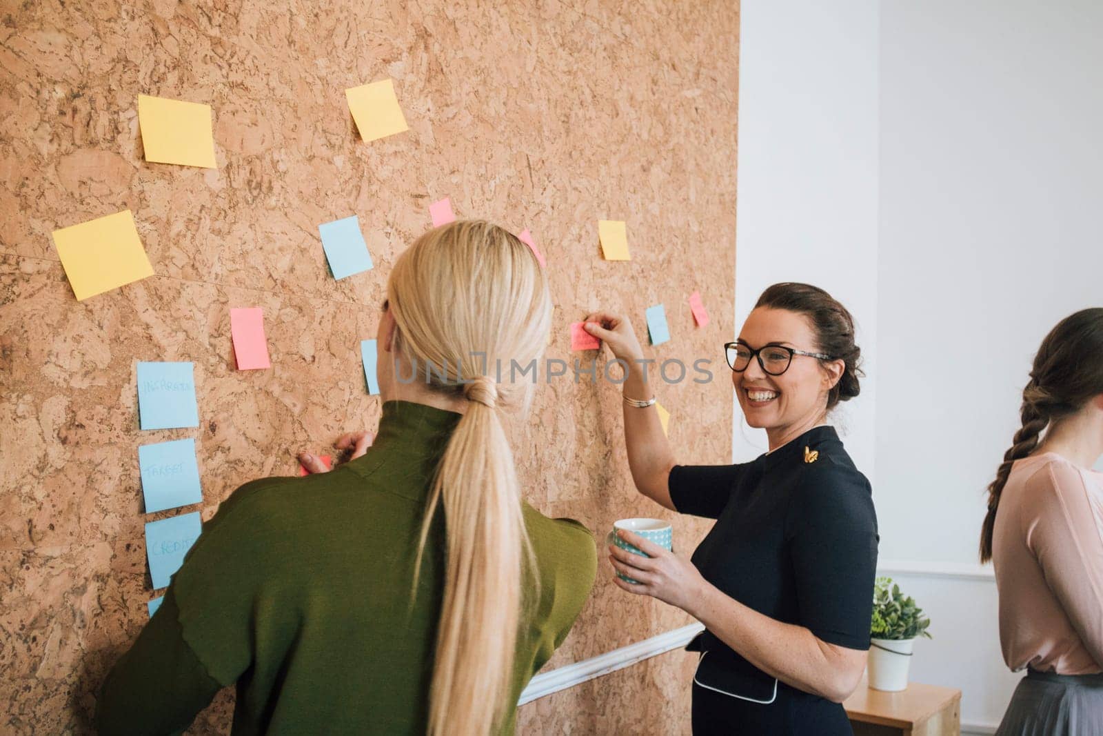 Two women are at work in an office. They are standing at a cork board and are having a discussion as they pin things up.