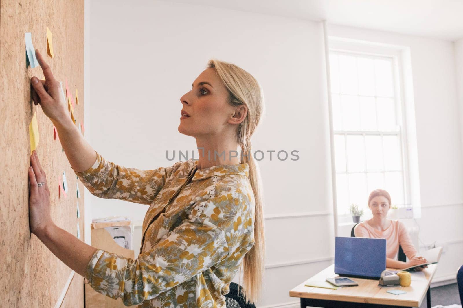 Female worker is pointing to a note on a cork board and is having a discussion with one of her coworkers, who is sitting at a desk in the background.