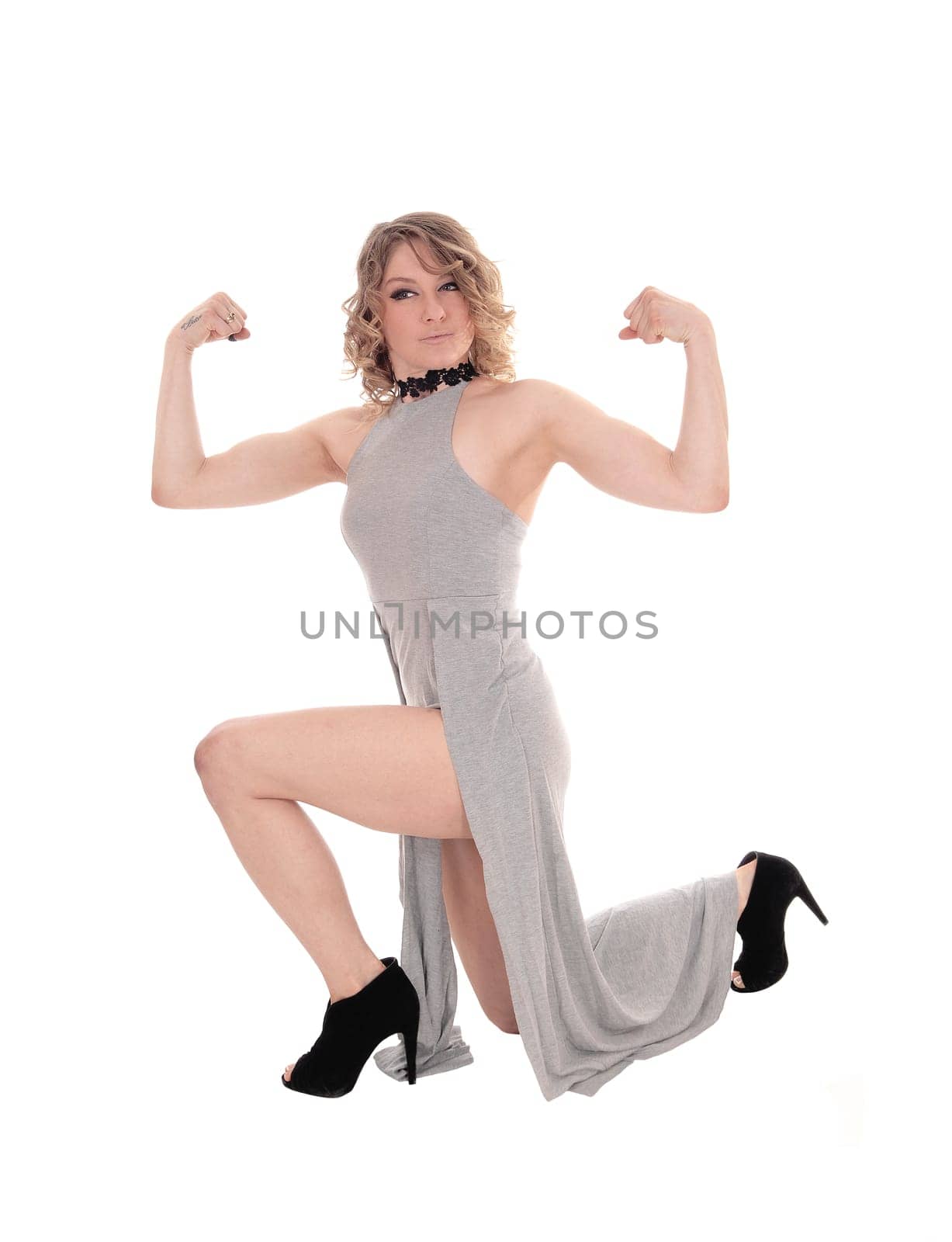 A beautiful blond woman kneeling on the floor in a gray dress and.flexing her muscles, isolated for white background..