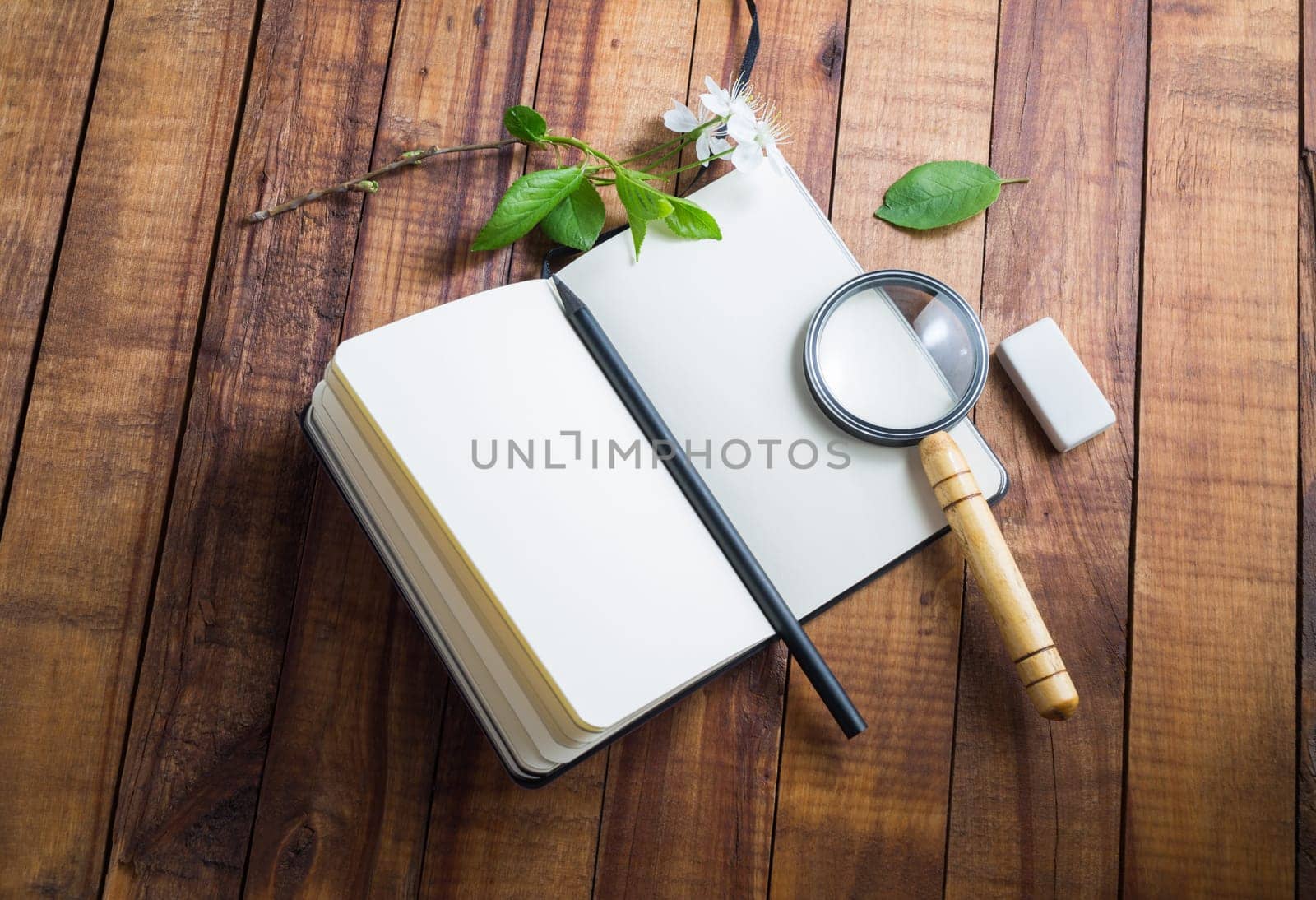 Photo of blank notebook, magnifier, pencil, cherry flowers and green leaves, eraser on vintage wood table background. Mockup for placing your design.