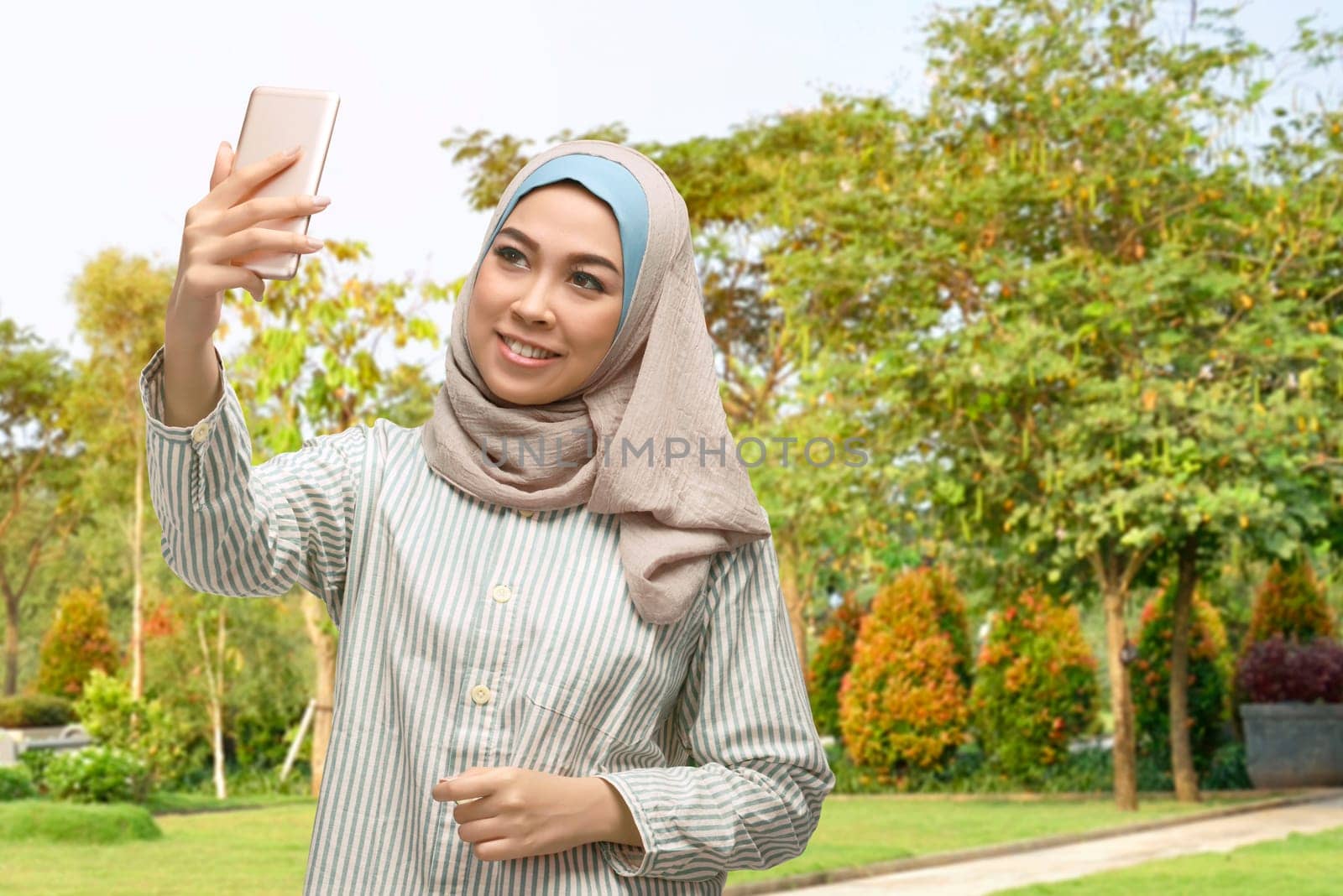 Beautiful asian muslim woman taking selfie photo with smartphone on the park