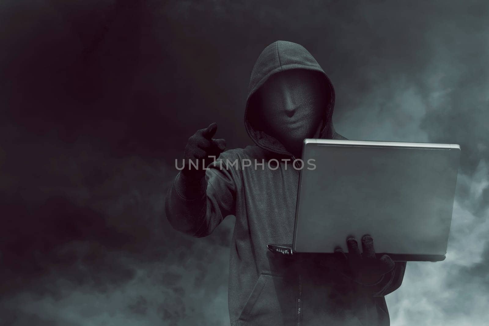 Portrait of hooded hacker with mask holding laptop while standing against dark room