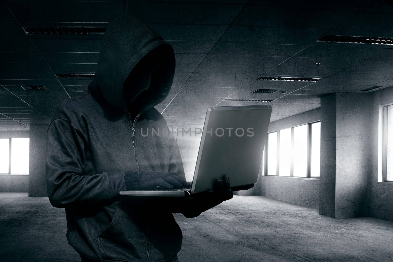 Hooded man with mask using laptop to hacking on the empty dark room