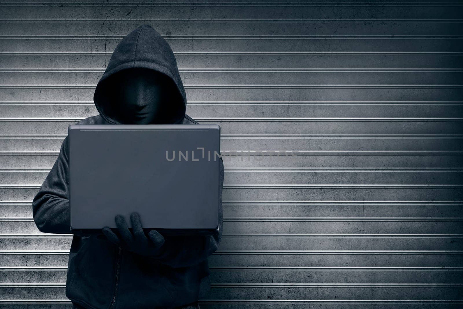 Hooded hacker with mask holding laptop while typing against metal roller shutter door background