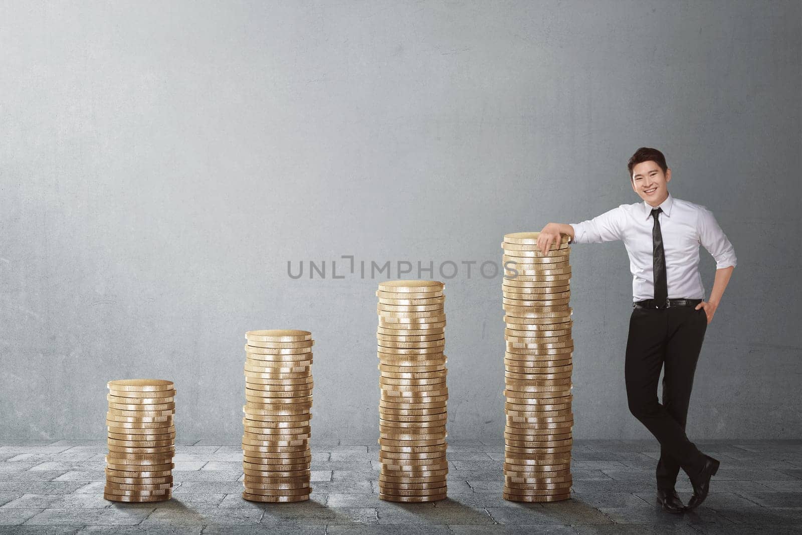Confident asian businessman standing beside coin stack against grey wall background