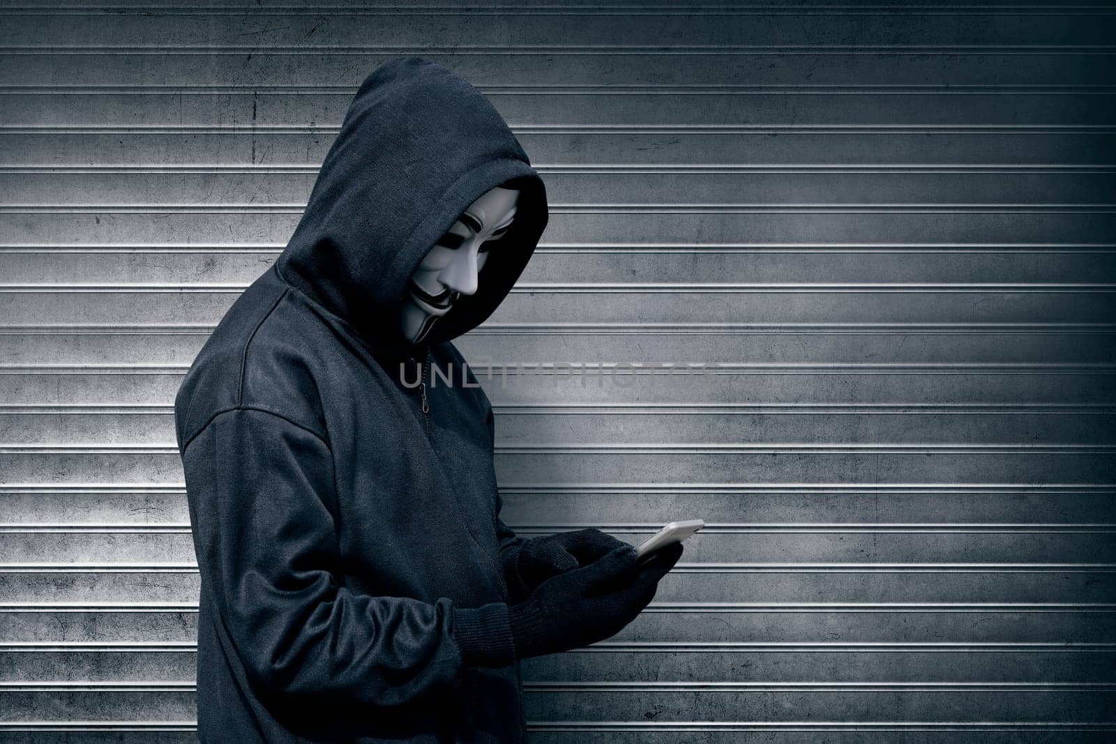 Hooded man with vendetta mask using mobile phone against roller shutter background