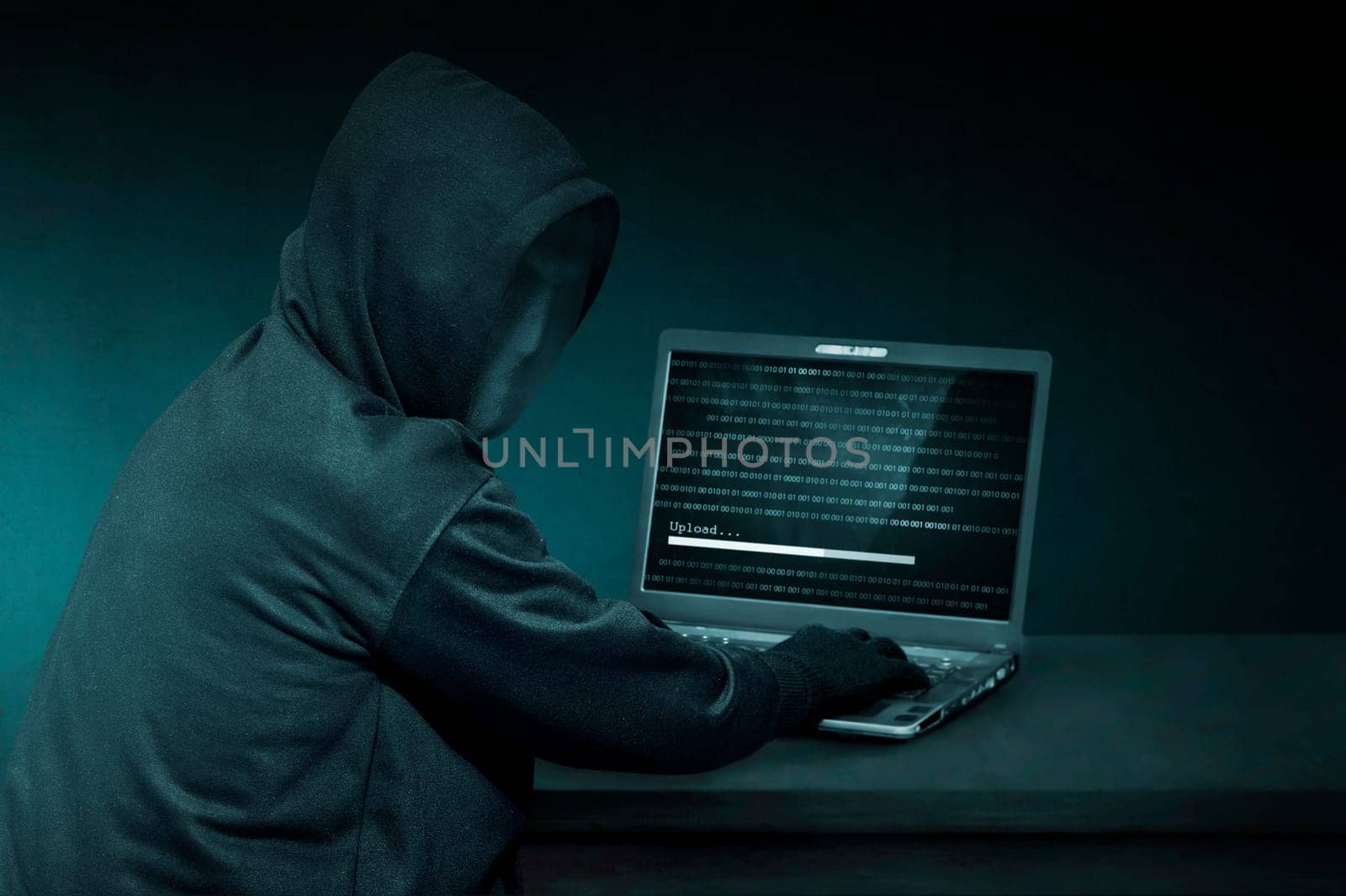 Hooded hacker with anonymous mask using laptop to steal data in the dark room