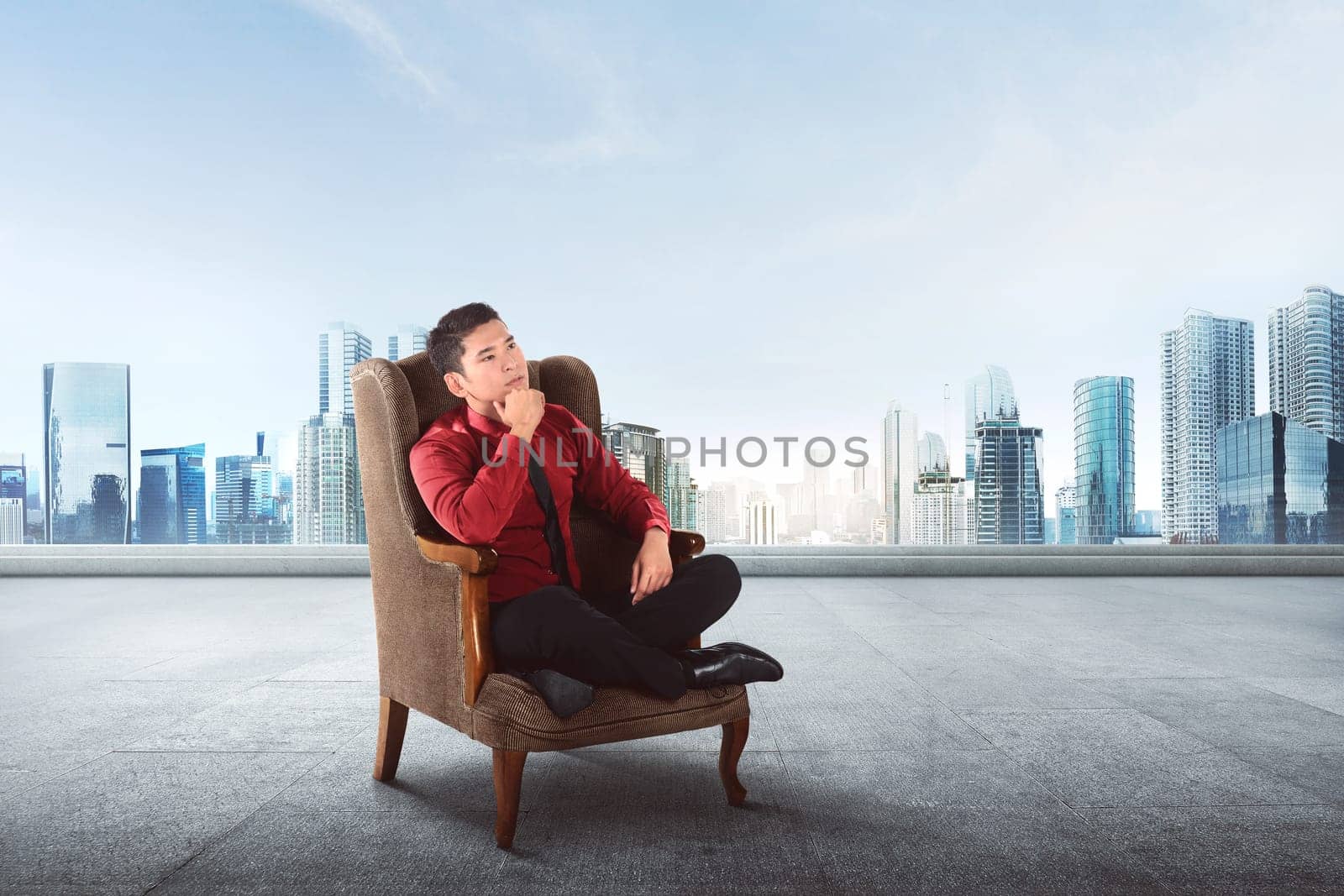 Thoughtful asian businessman sitting on sofa thinking about something with skyscraper background