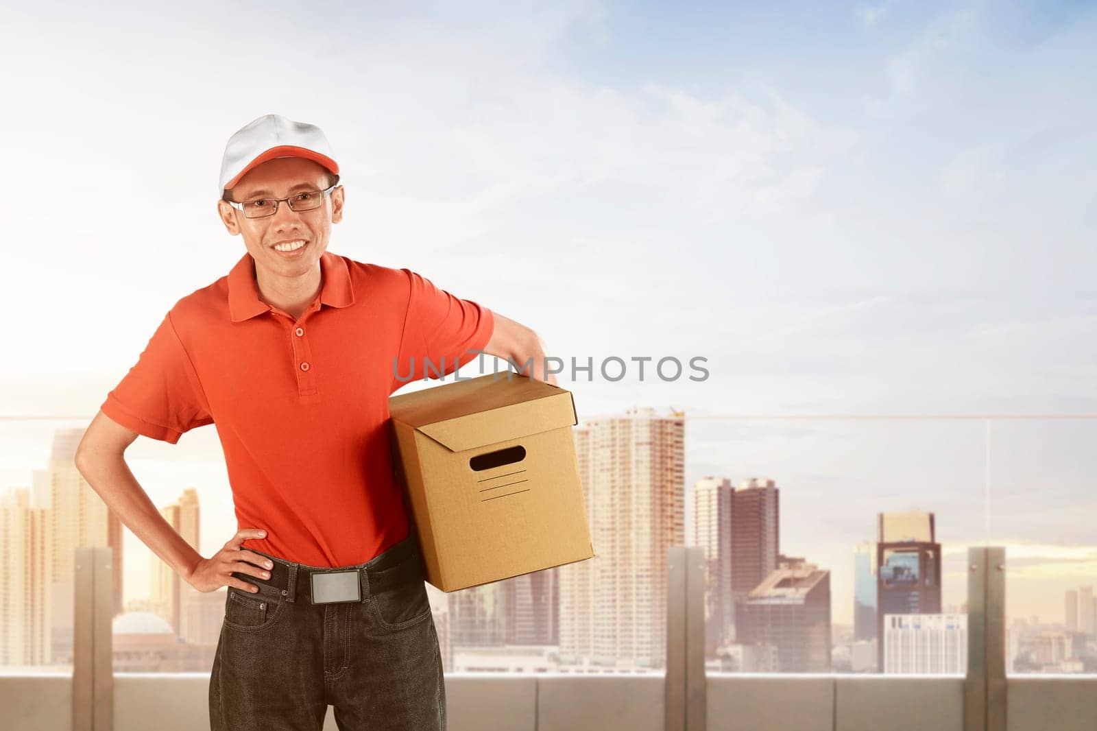 Happy asian courier holding big parcel against cityscape background