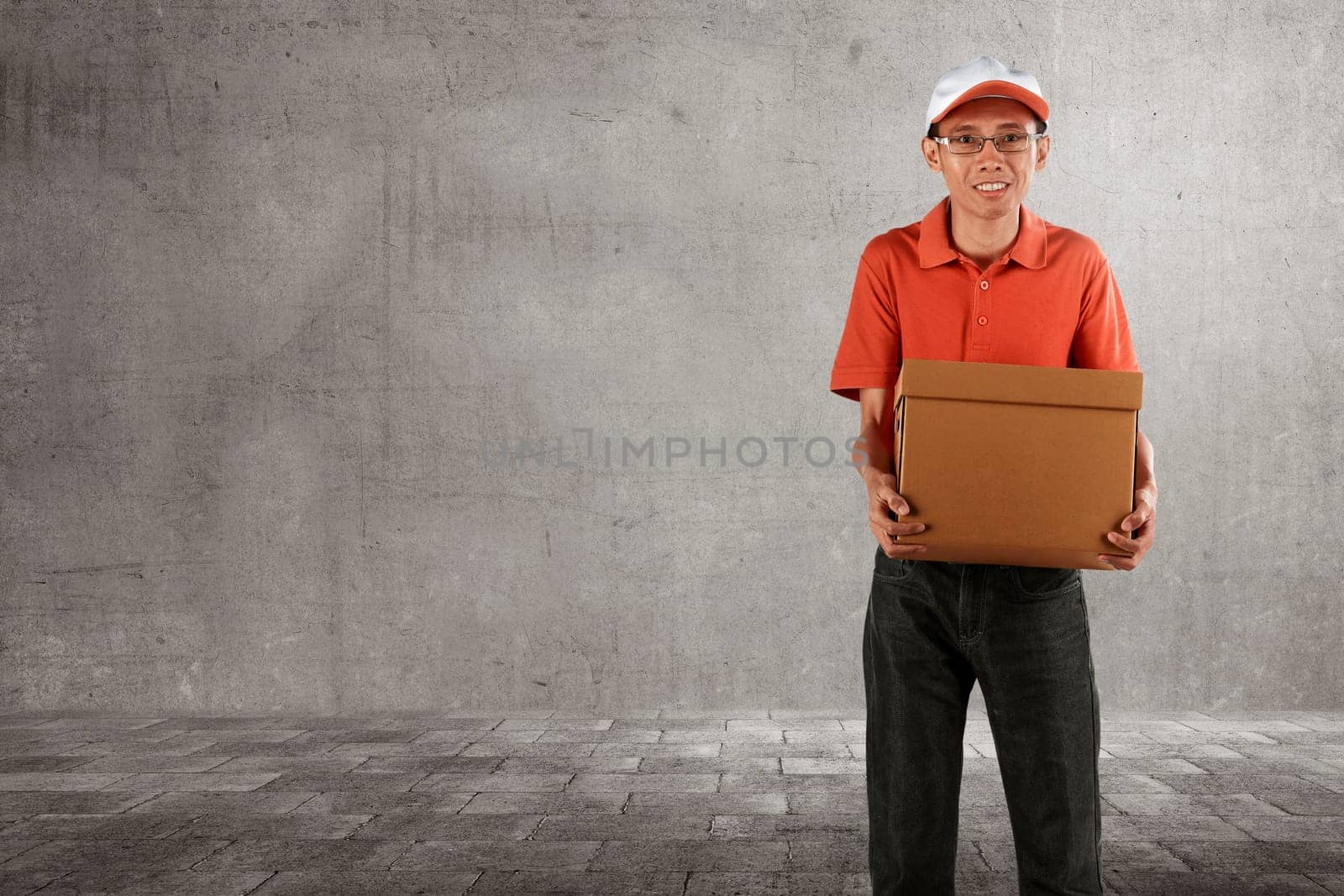 Cheerful asian courier standing with parcel over grey wall background