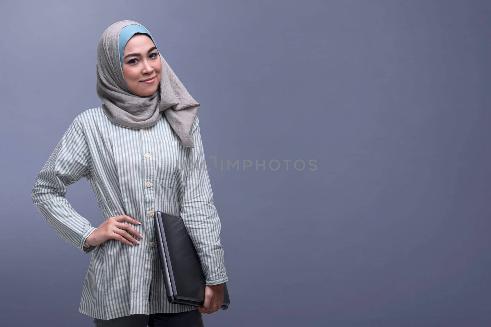 Happy asian muslim woman wearing veil holding laptop against dark background