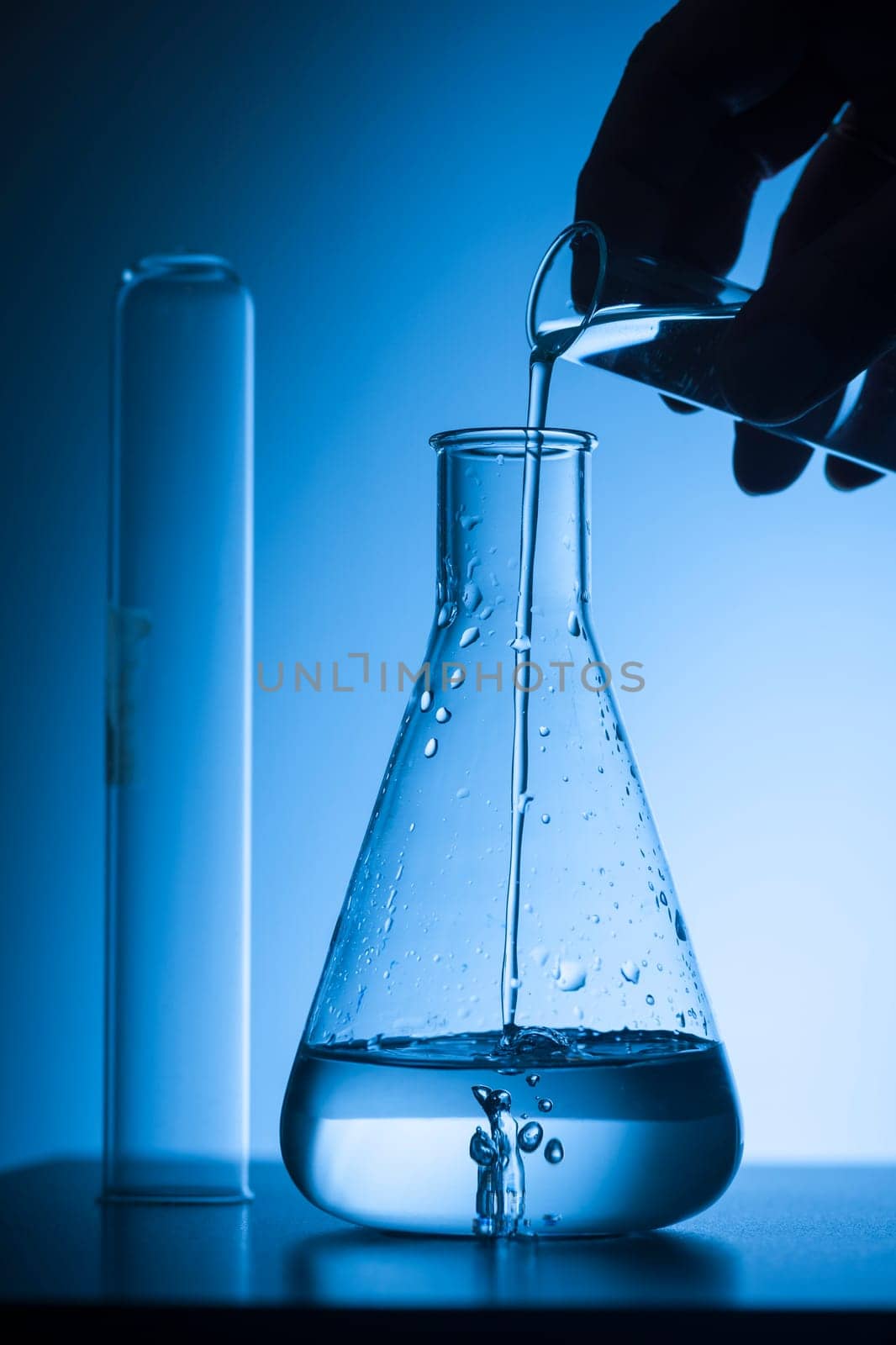 Hand sihouettes with test tube fills an Erlmeyer flask against a blue tinted studio background.
