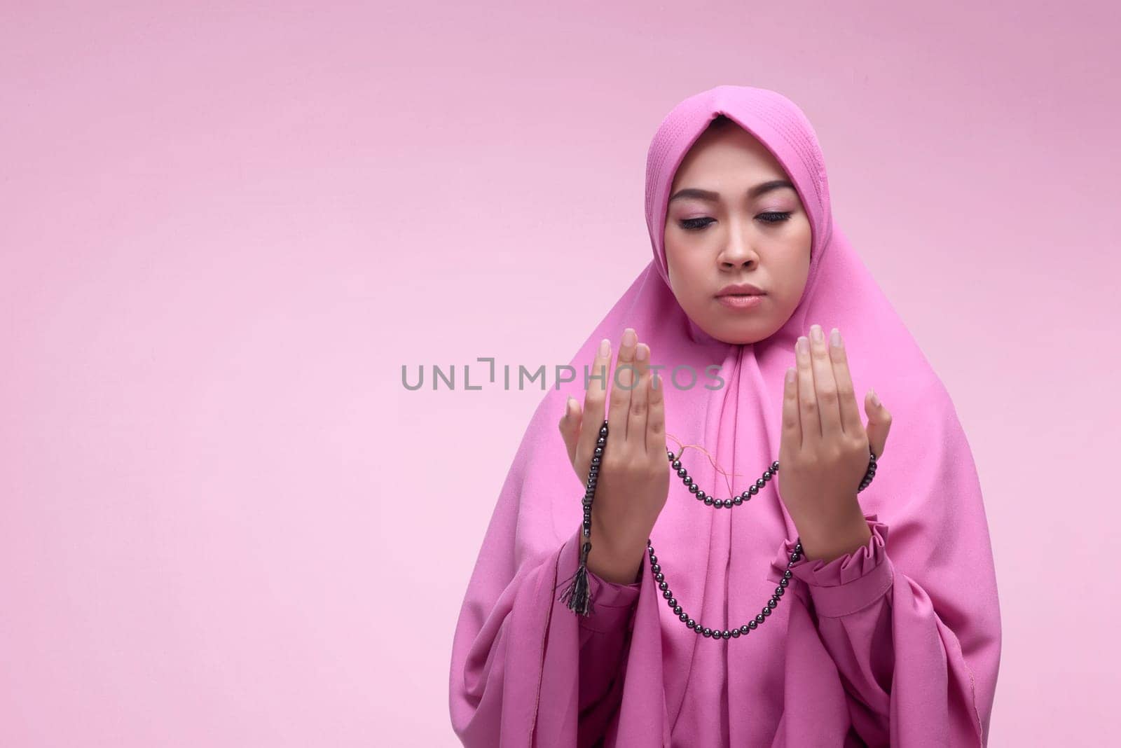 Young asian muslim woman praying with prayer beads over pink background