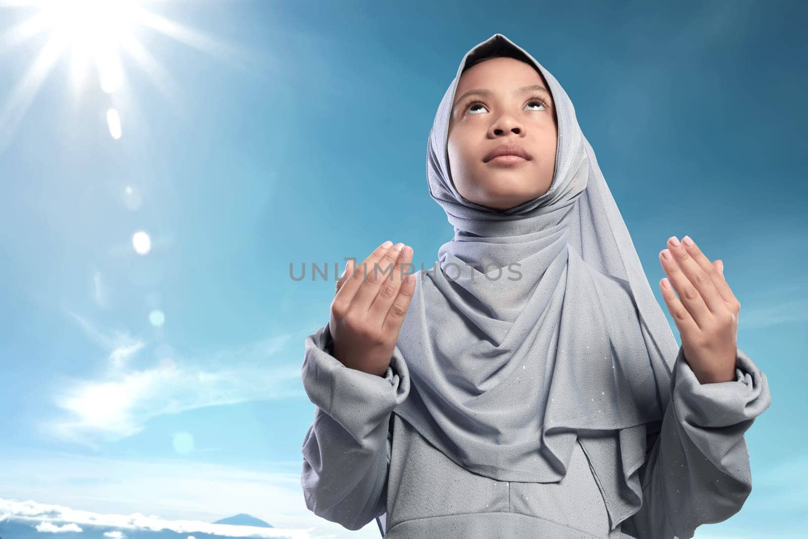 Religious asian muslim kid raising hand and praying against blue sky background