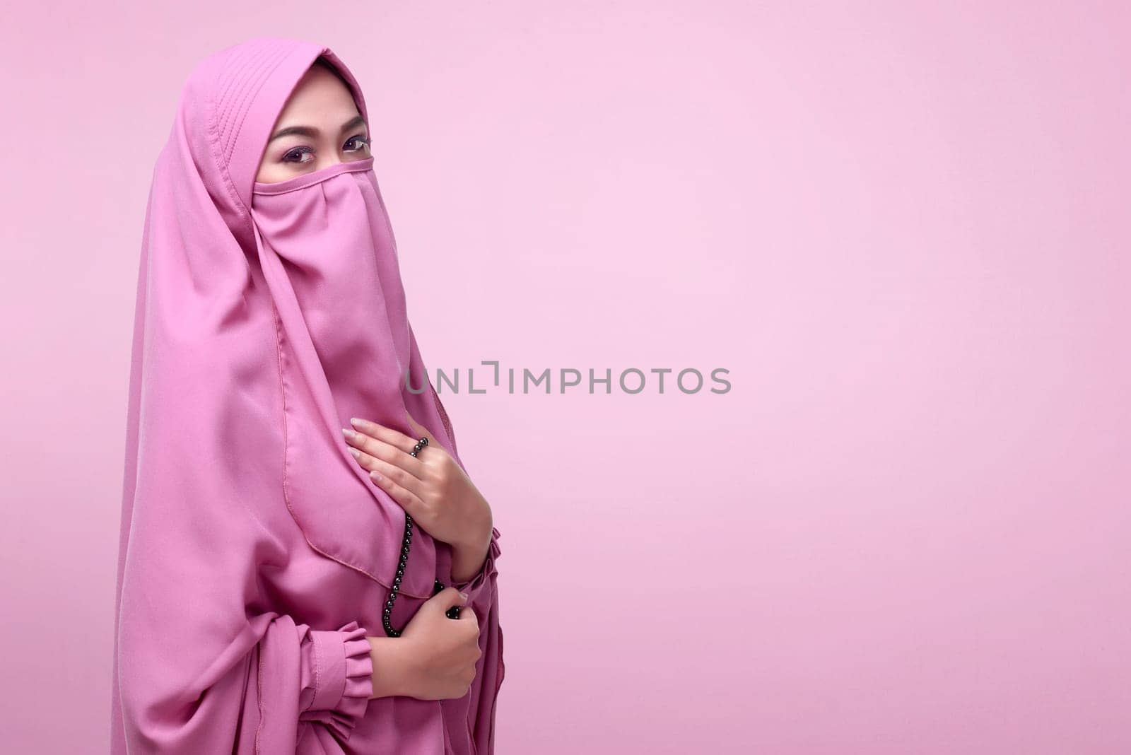 Young asian muslim woman in veil with prayer beads standing over pink background