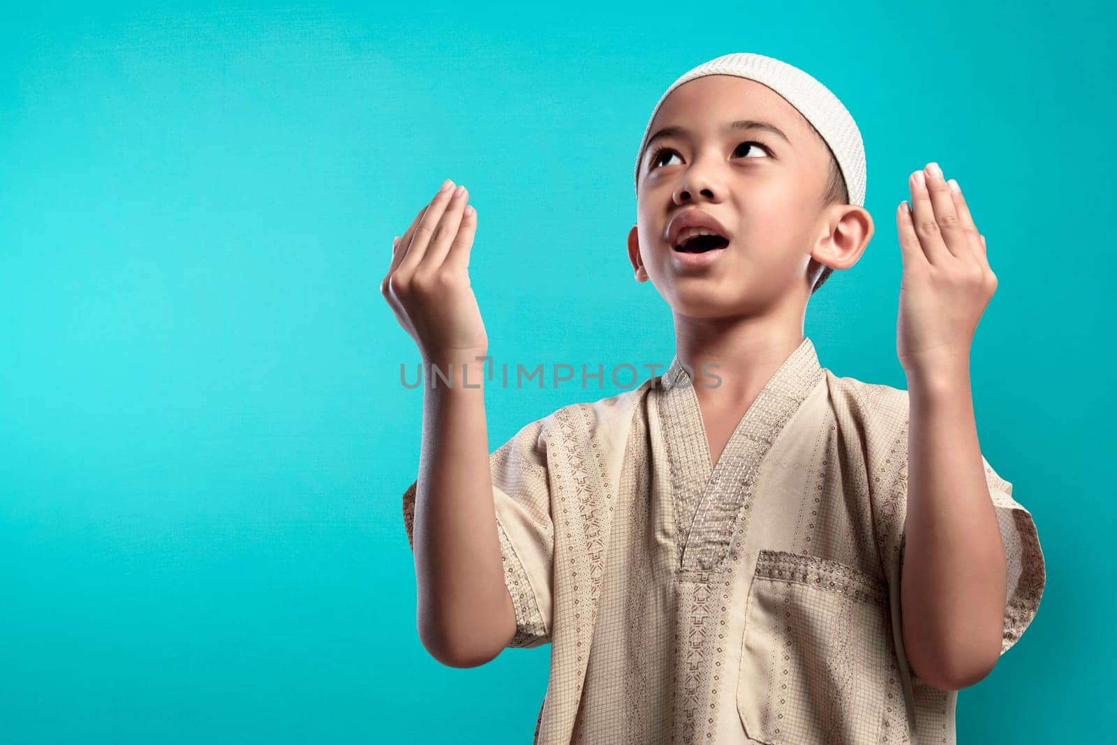 Little asian muslim kid raising hand and praying to god over blue background