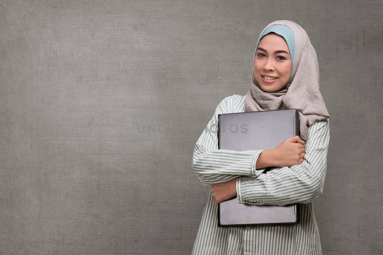 Beautiful asian muslim woman holding laptop against concrete background