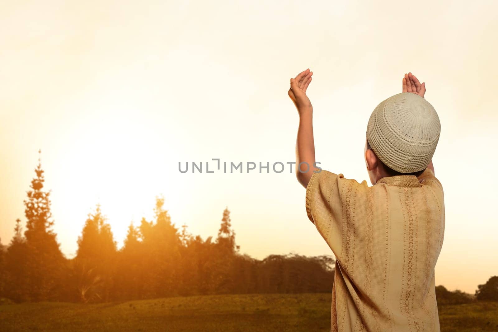 Back view of asian muslim kid with cap praying to god in nature during sunset